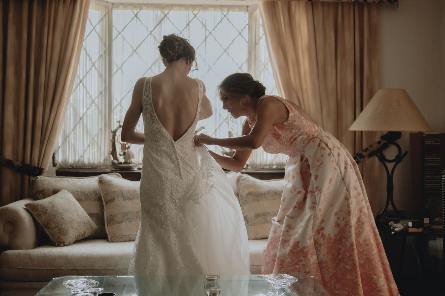 Bridesmaid helping bride with final wedding dress adjustments in sun-lit room.