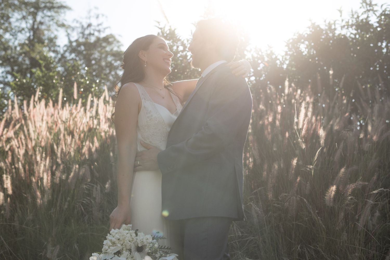Wedding couple embraces in golden sunset light amidst tall grasses.