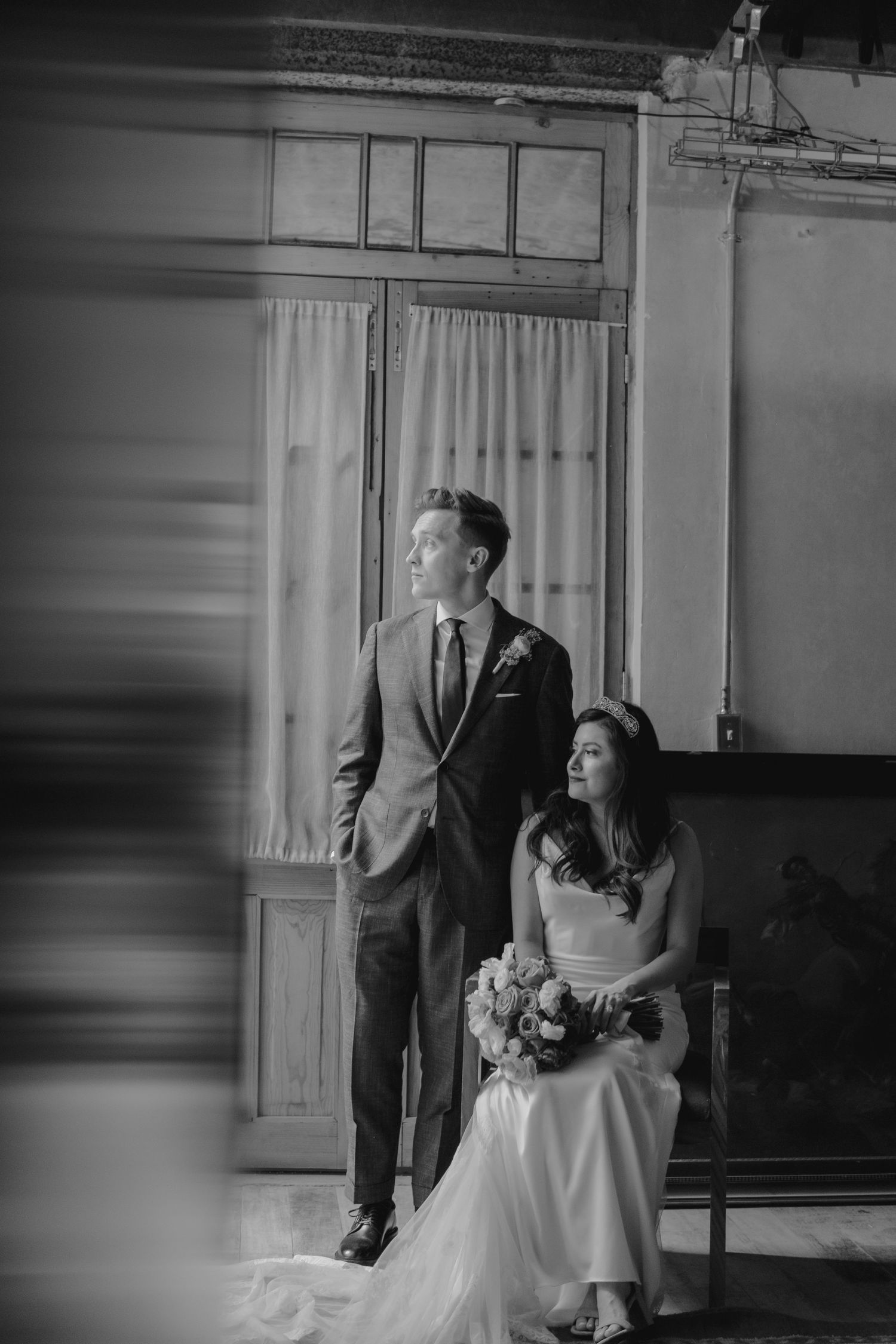 Artistic wedding portrait showing couple in formal attire seated by vintage windows with natural light.
