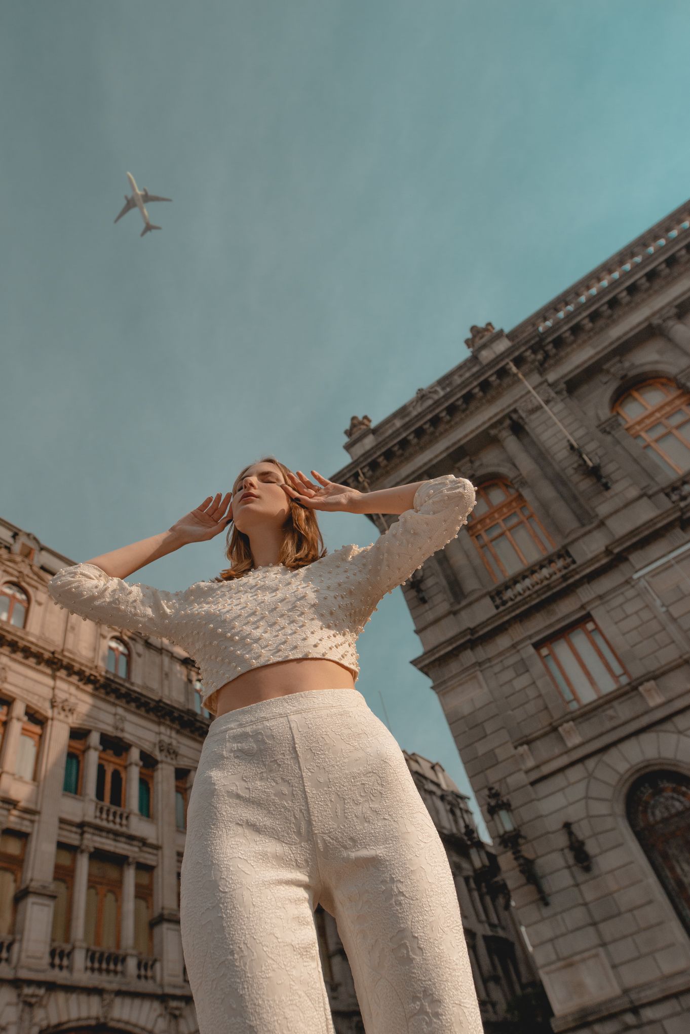 Fashion pose against classic architecture with airplane in sky and turquoise backdrop.