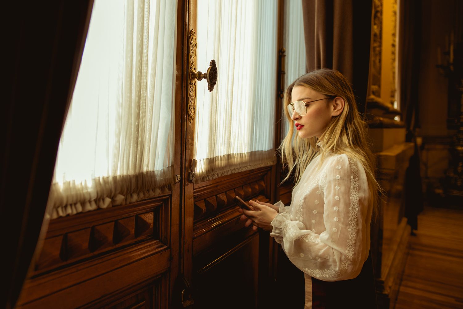 Person in white Victorian blouse looking through ornate window in vintage interior setting.