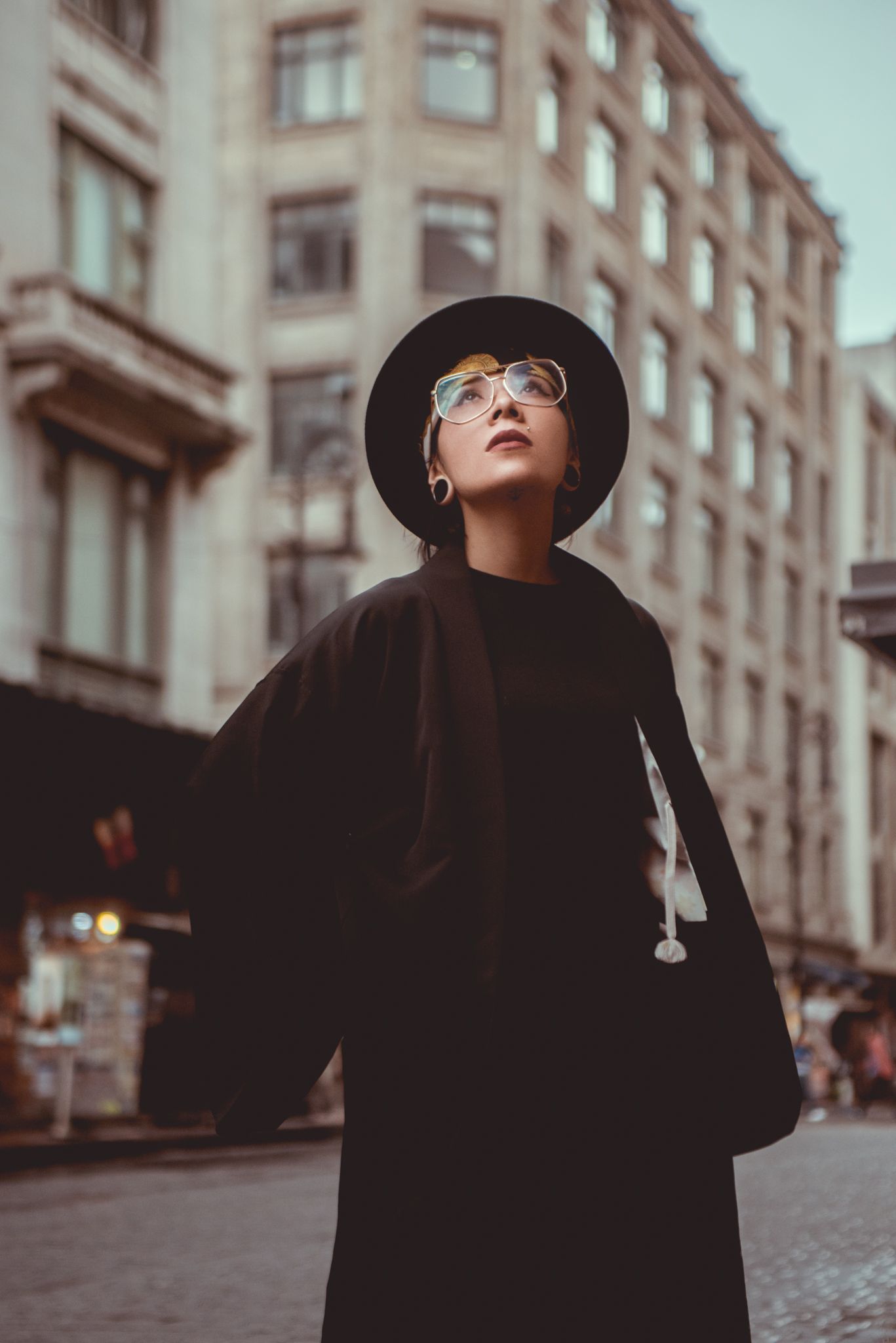 Person in black coat and hat looking upward on city street.