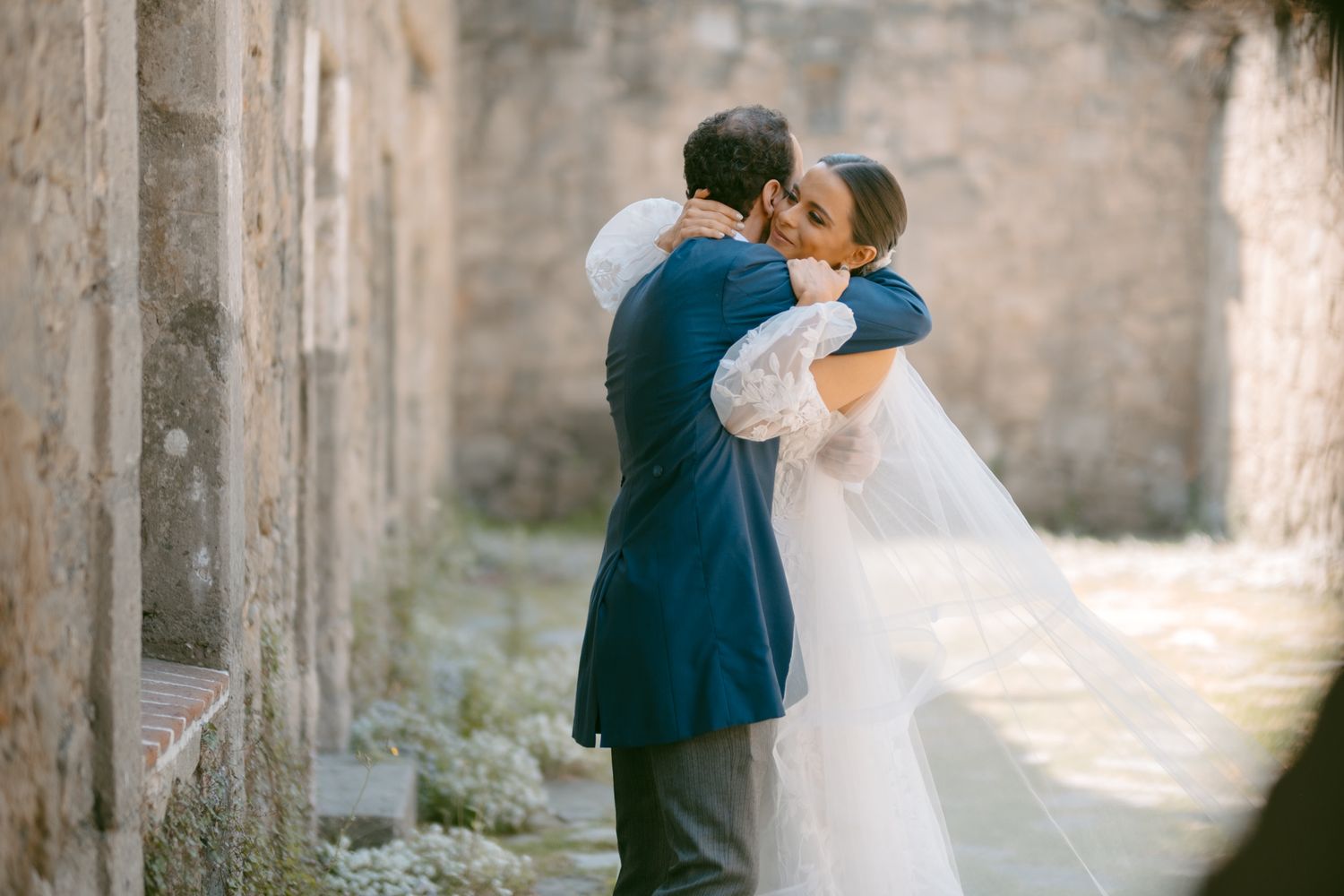 A romantic embrace between newlyweds against an ancient stone wall backdrop.