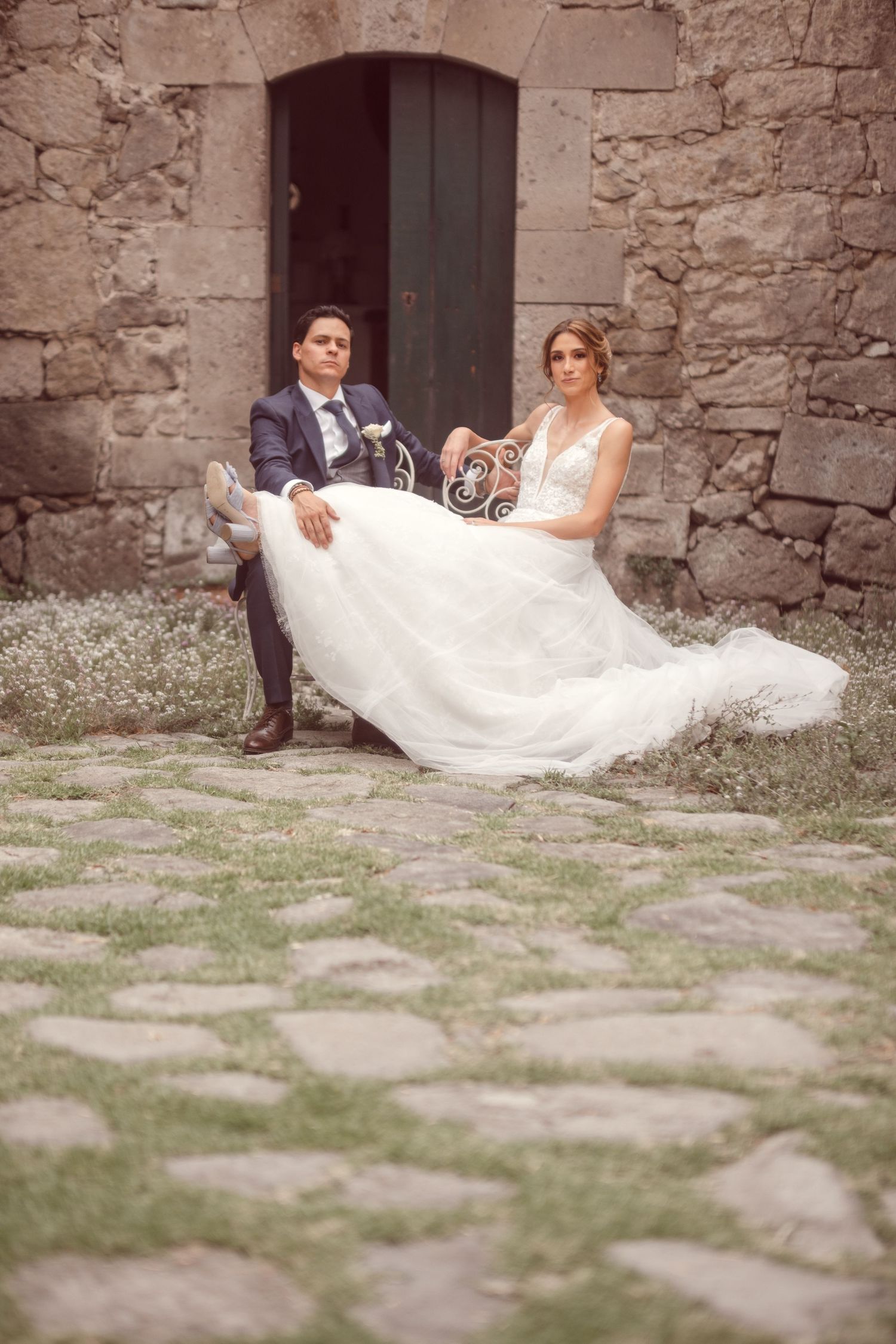 A couple in wedding attire sits on stone steps against a rustic wall with an arched doorway.