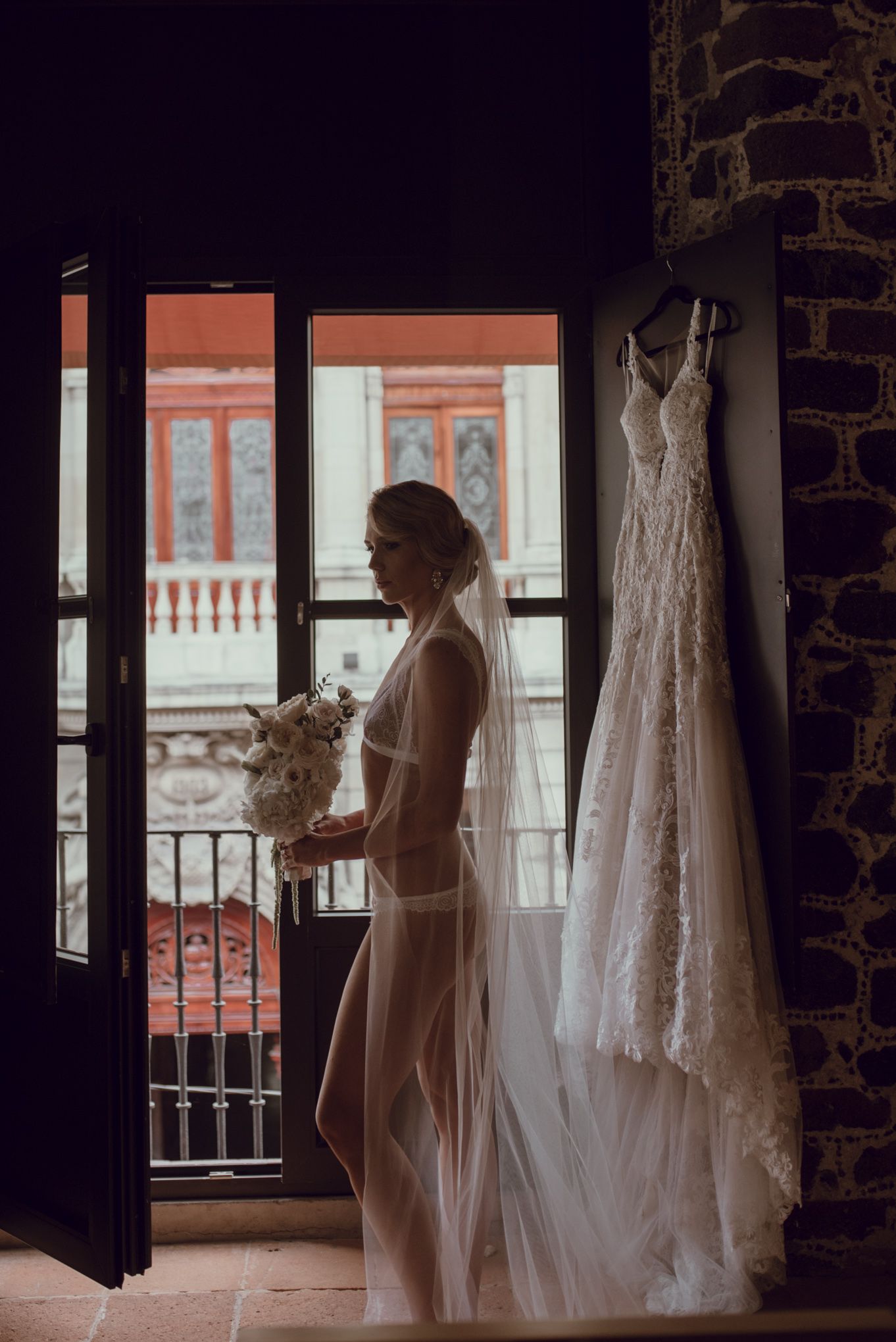 Silhouette of bride by balcony window with wedding dress hanging nearby.