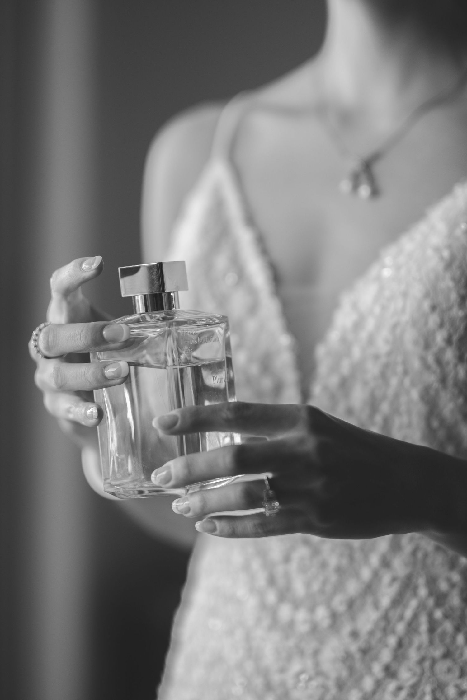 Delicate hands holding a crystal perfume bottle against a beaded wedding dress in black and white.