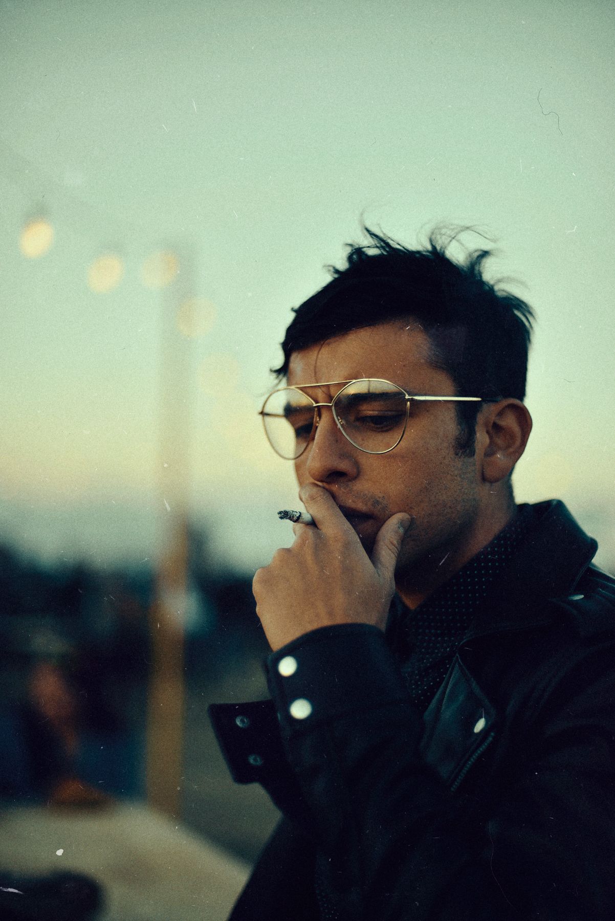 Moody portrait of man in glasses smoking cigarette at dusk.