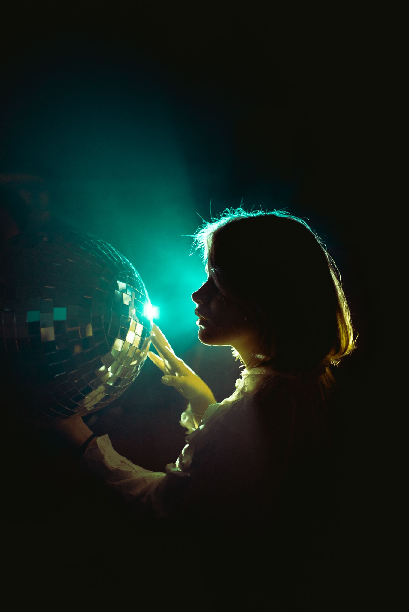 Silhouette holding disco ball illuminated by turquoise backlighting in dark setting.