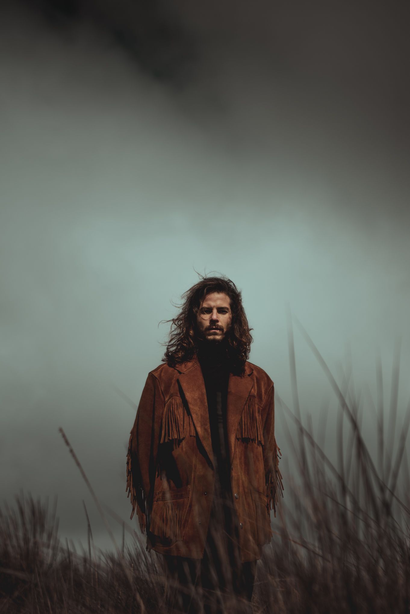 Figure in brown fringe jacket standing in misty field at dusk.