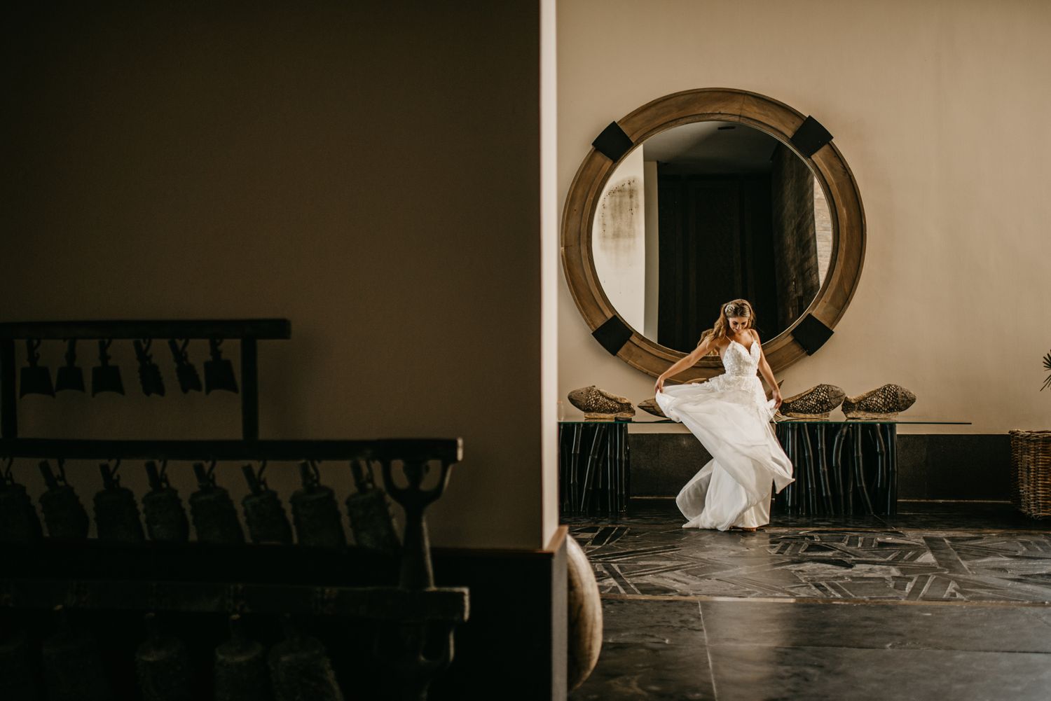 Someone in a flowing white wedding dress sits elegantly in front of a large round mirror in a minimalist interior space.