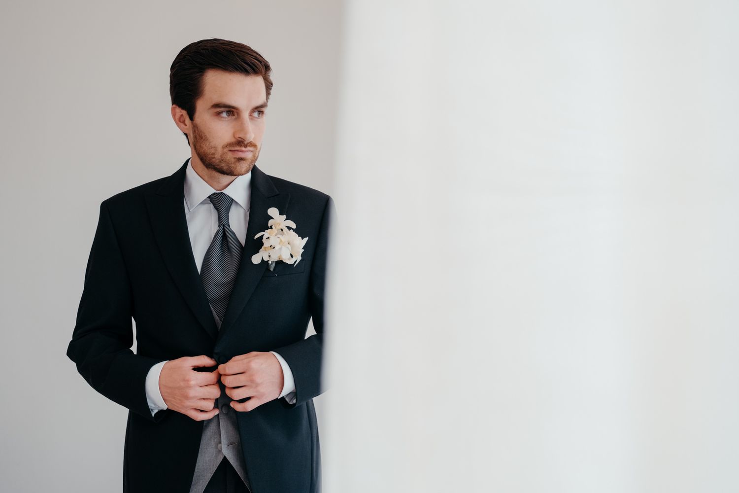 Groom wearing classic black tuxedo with boutonniere adjusts jacket buttons against light wall.