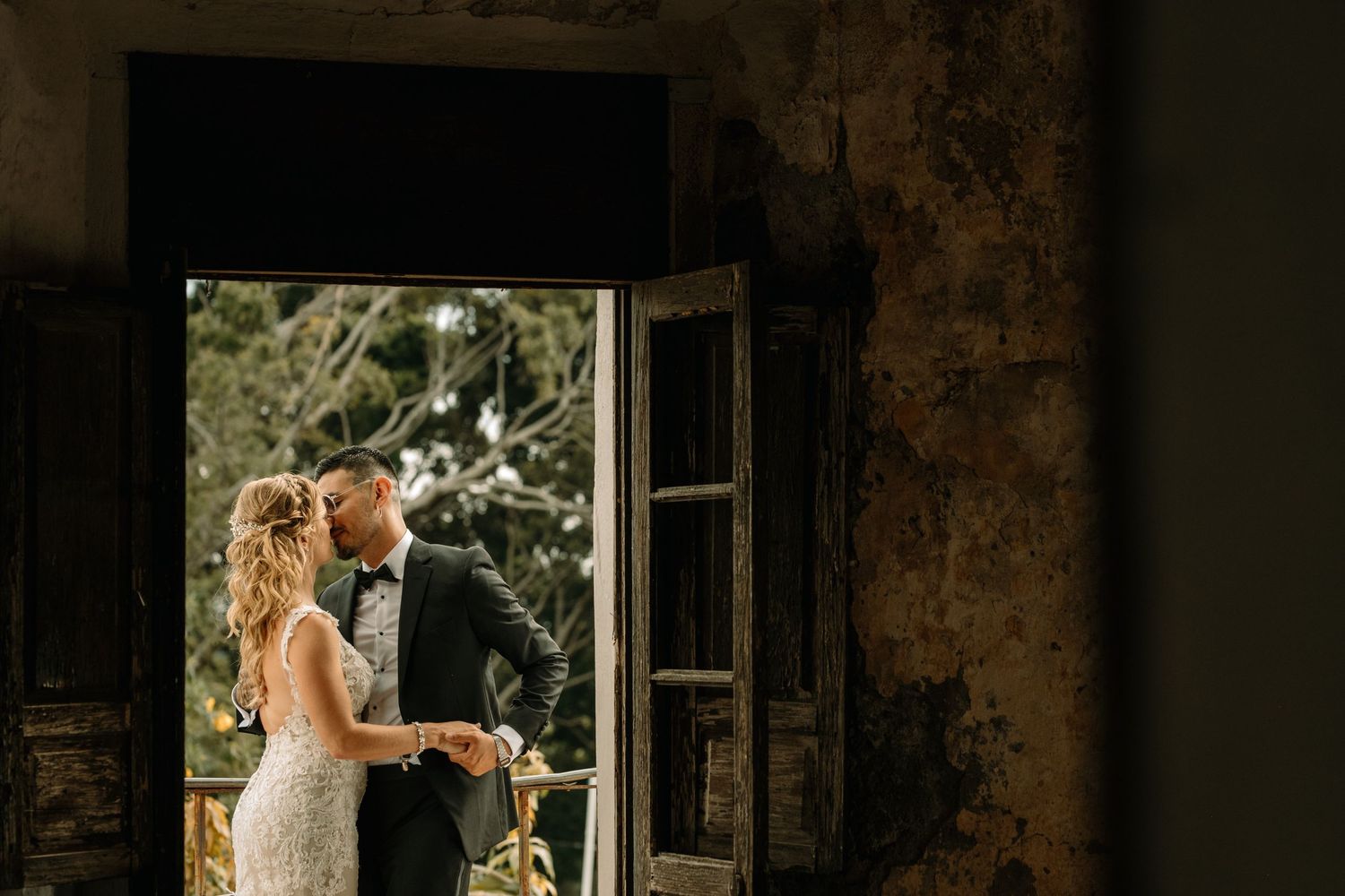 A couple in wedding attire stand together in golden light by a rustic open doorway.