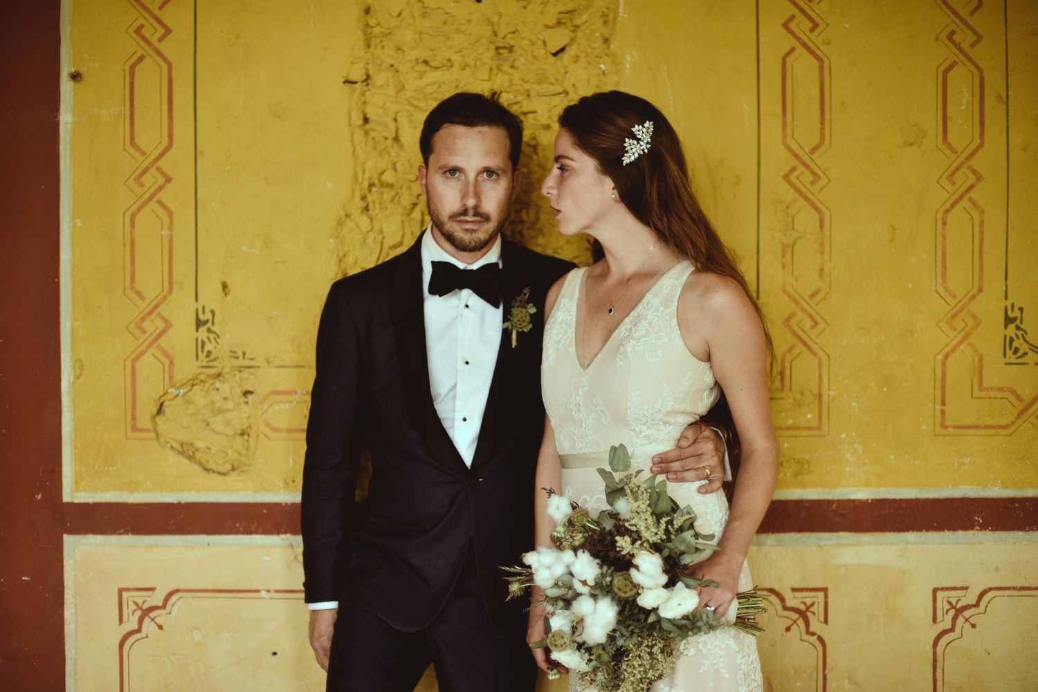 A couple in formal wedding attire poses against a yellow wall with decorative red trim during their classical ceremony.