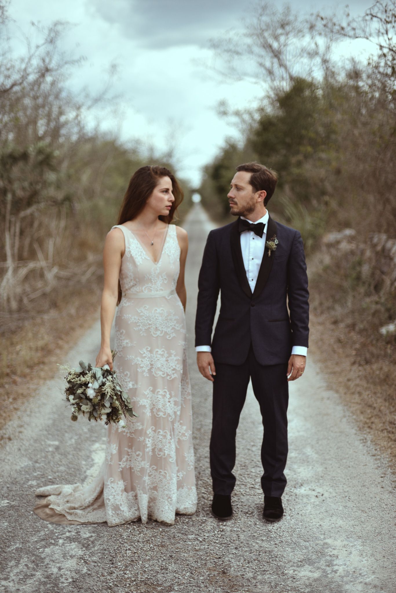 Couple stands together on forest path with bride wearing lace gown holding bouquet and groom in classic tuxedo.