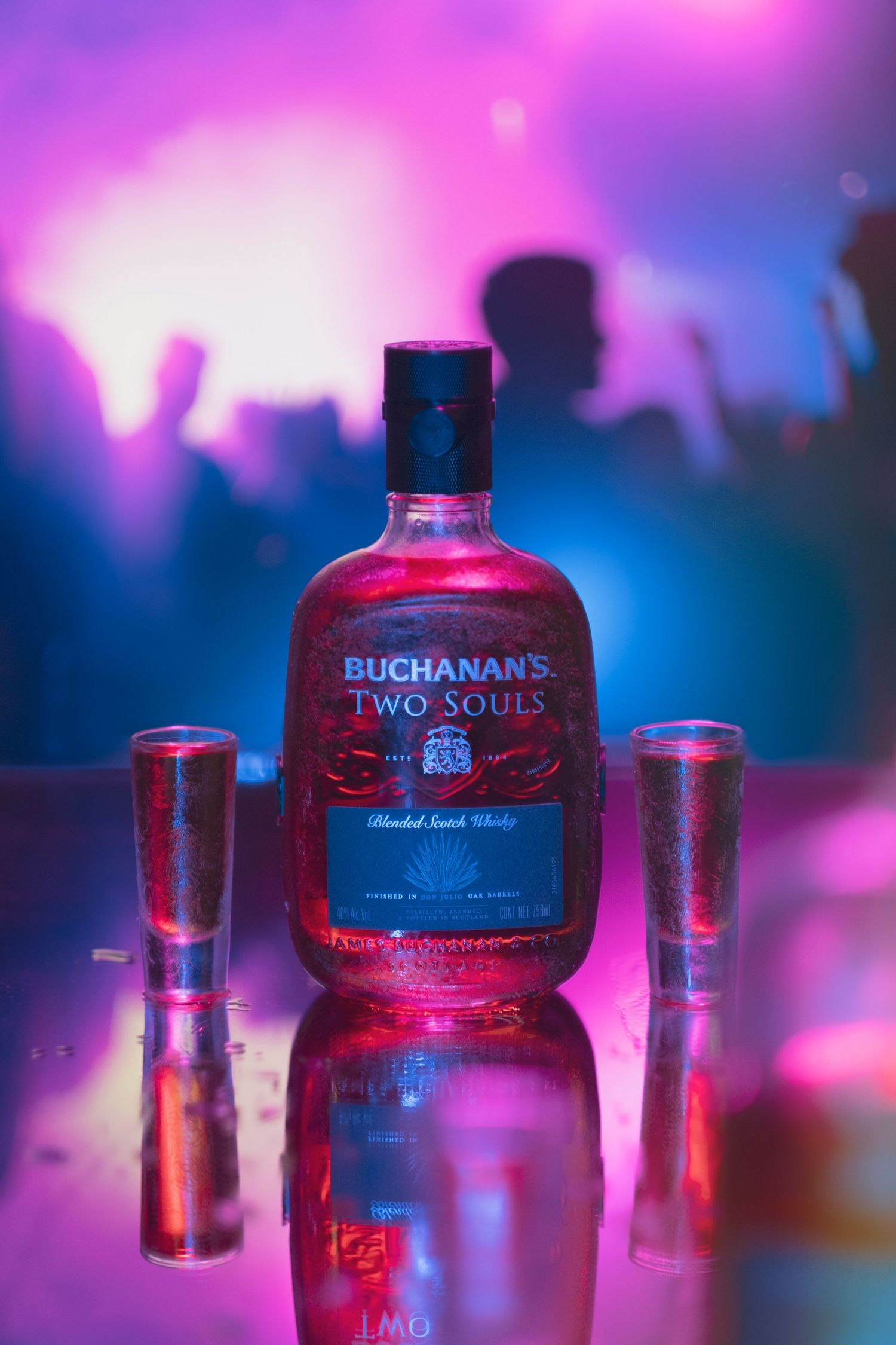 Several bottles and glasses of Two Souls liquor illuminated by colorful pink and blue lighting in a nightclub setting.