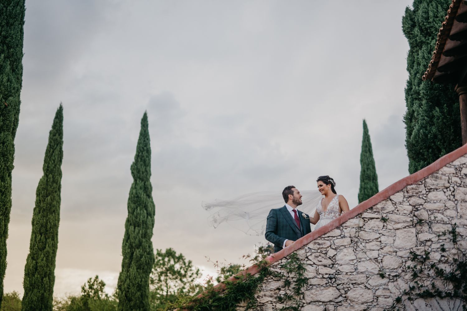 A couple in formal attire sits together on stone steps with tall cypress trees and moody sky in the background.