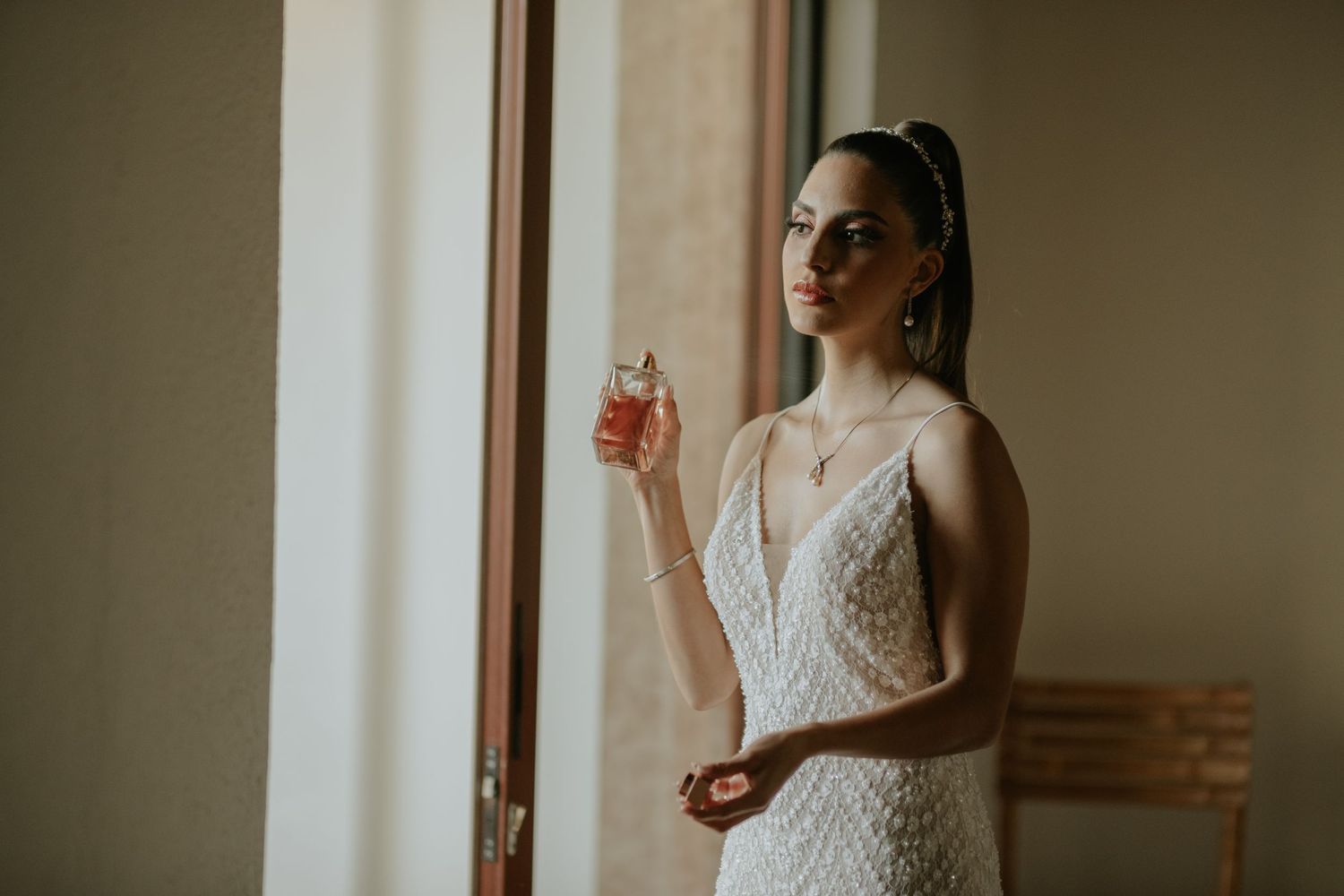 A person in an elegant white lace wedding dress holds a champagne glass while standing by a window in soft lighting.