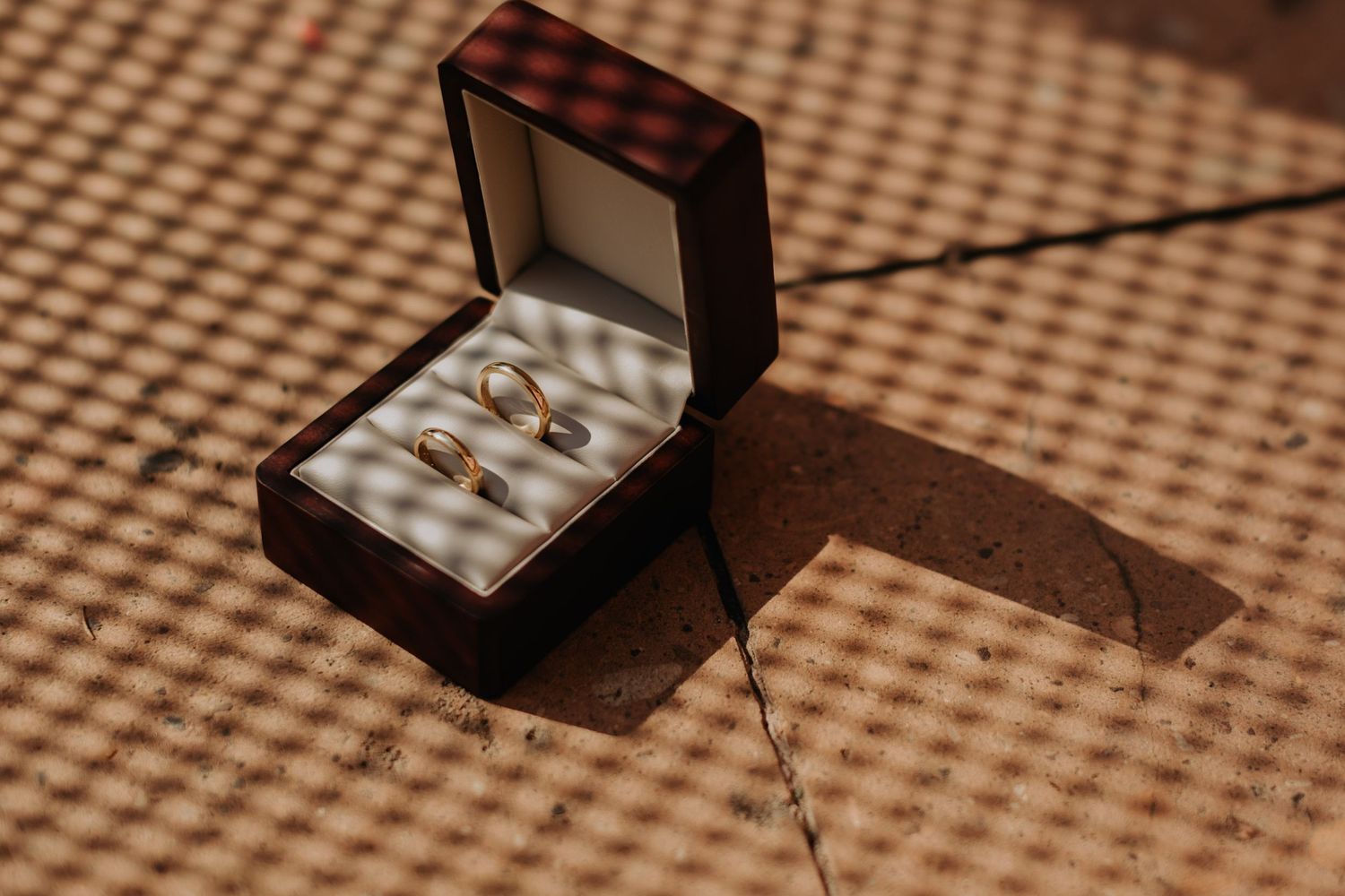 Wedding rings nestled in a wooden jewelry box casting dramatic shadows on textured fabric background.