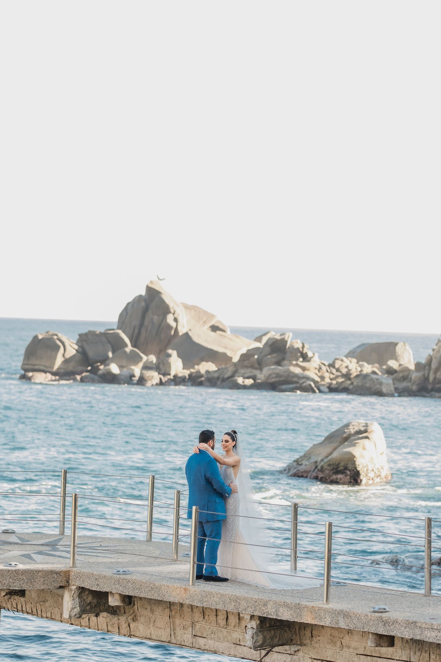 A couple embraces on a seaside pier with large rocky formations and blue Mediterranean waters in the background.