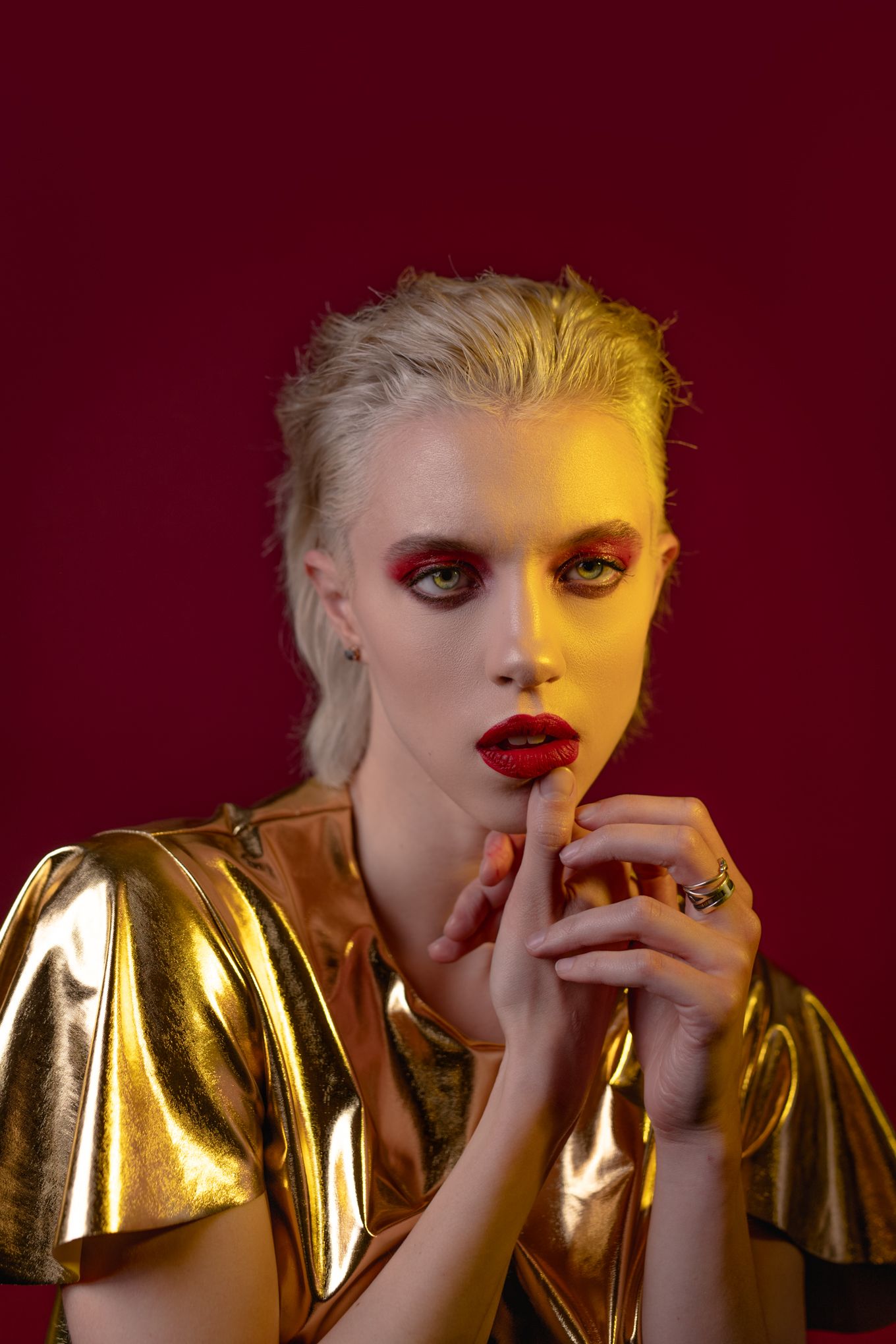 Fashion portrait with gold metallic top against burgundy background with dramatic makeup and lighting.