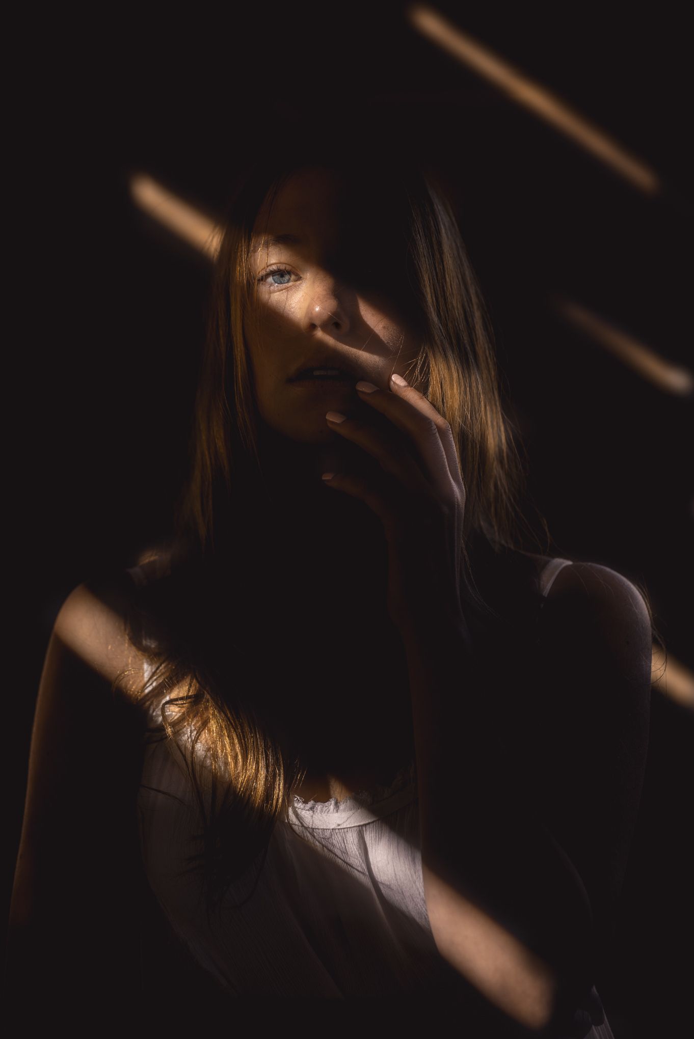 Dramatic portrait with light beams creating shadows across a face in dark moody lighting.