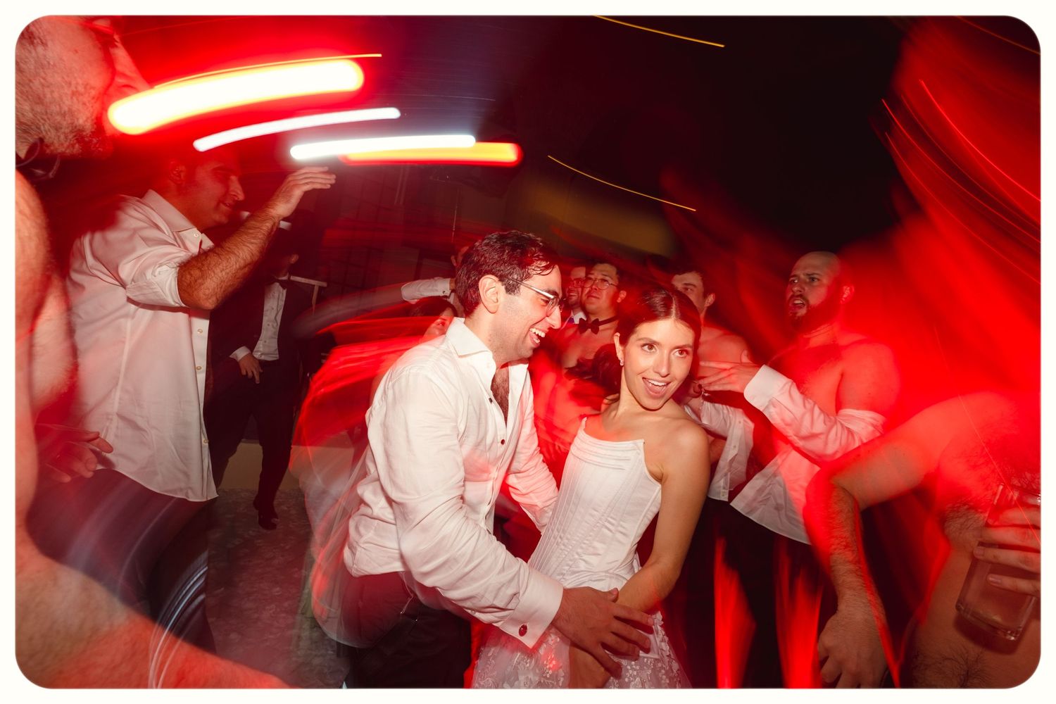Wedding party dancing energetically under red party lights on a dance floor at reception.