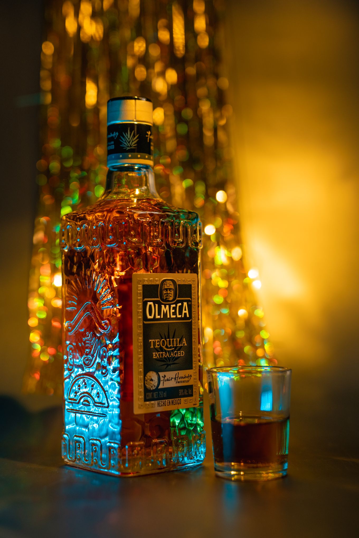 Olmeca tequila bottle glowing with colorful ambient lighting and shot glass on dark background.