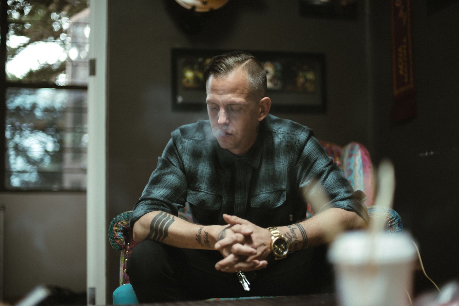 Person in plaid shirt sits thoughtfully in dimly lit room with decorative elements on wall.