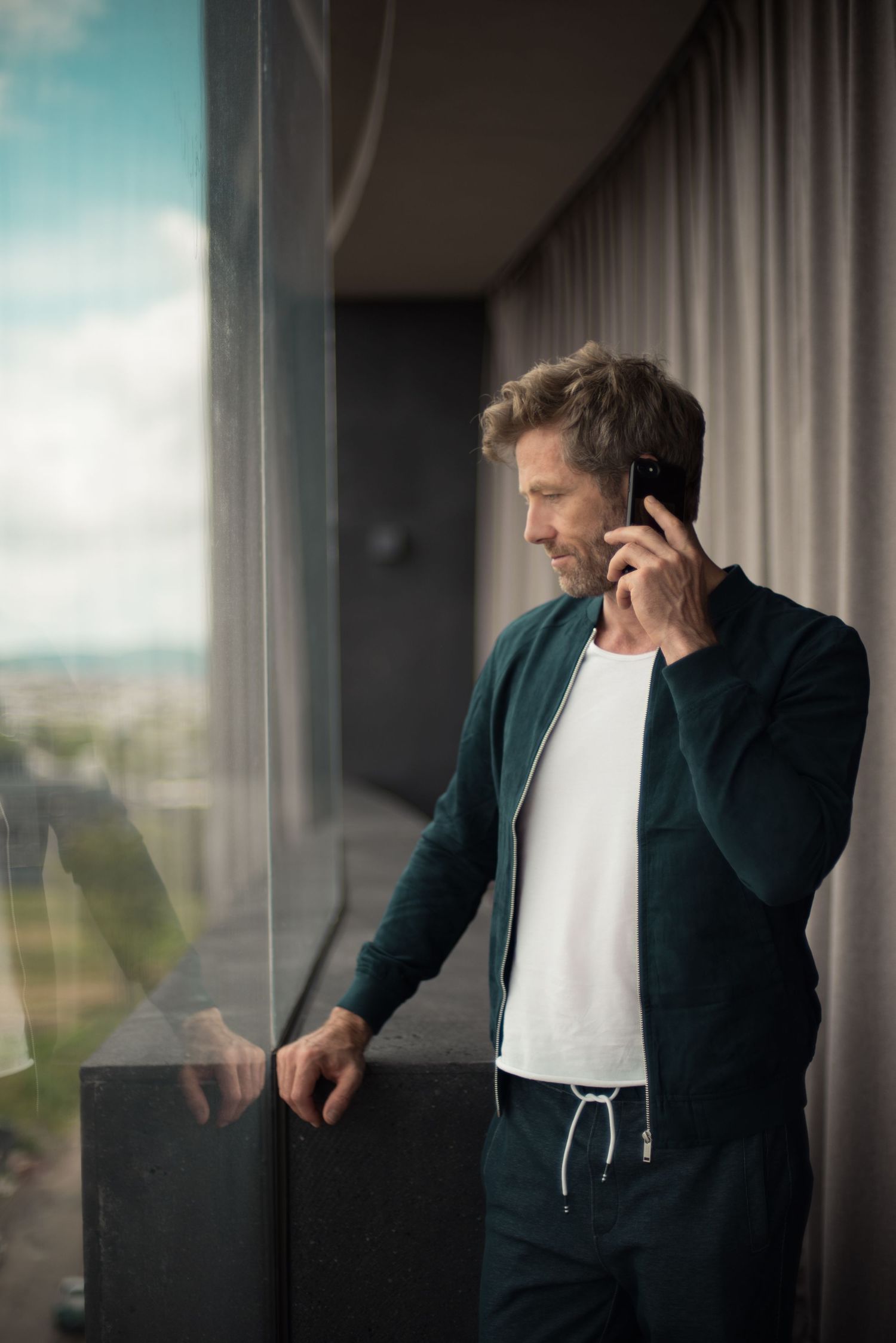 Man in dark jacket looking out window while on phone call.