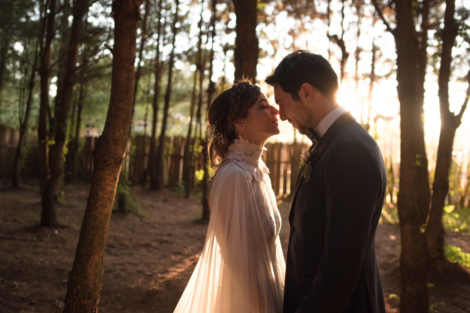 Romantic sunset portrait of a couple in elegant wedding attire standing close together in a pine forest.