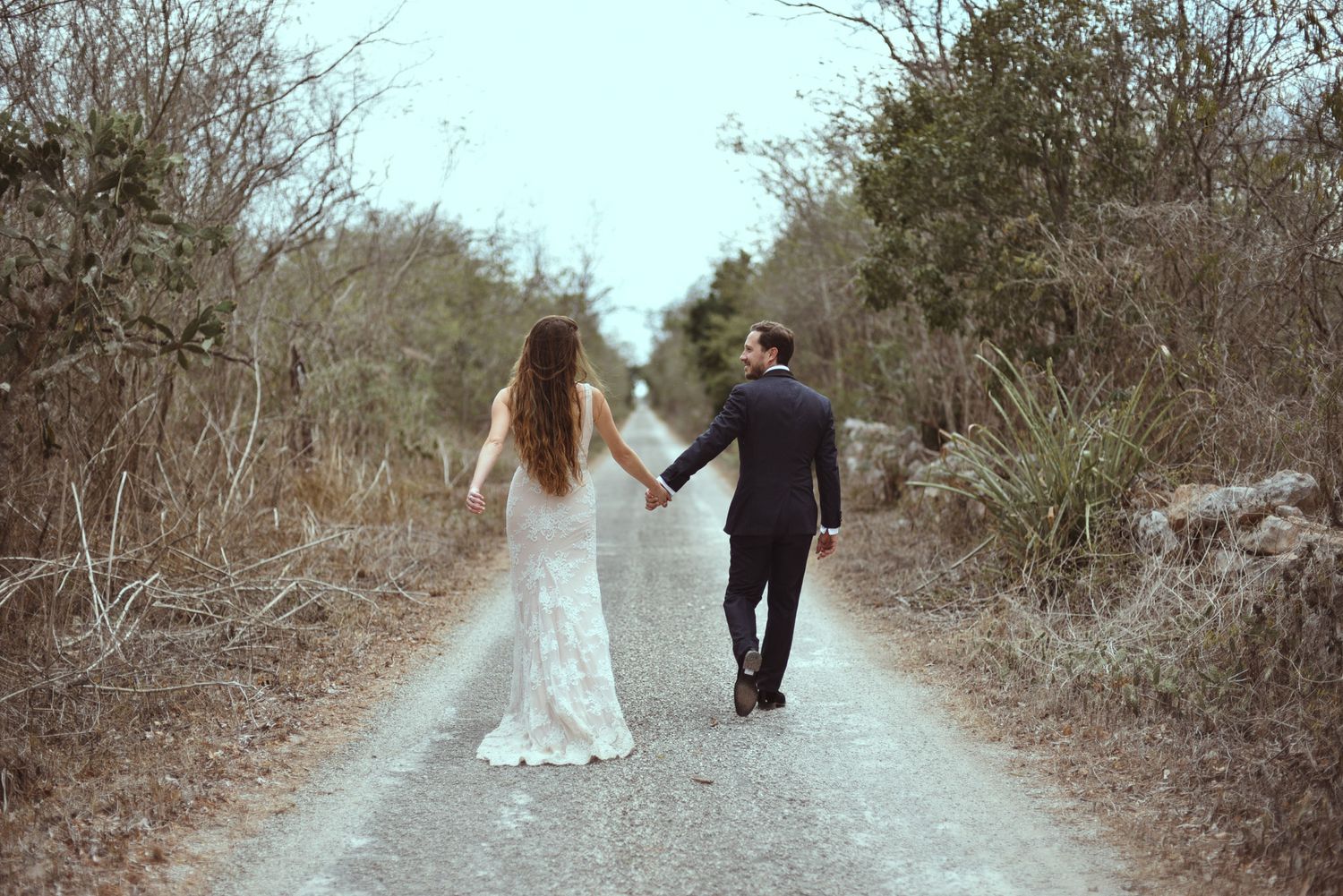 A couple in wedding attire walking hand in hand down a rustic dirt path surrounded by brush.