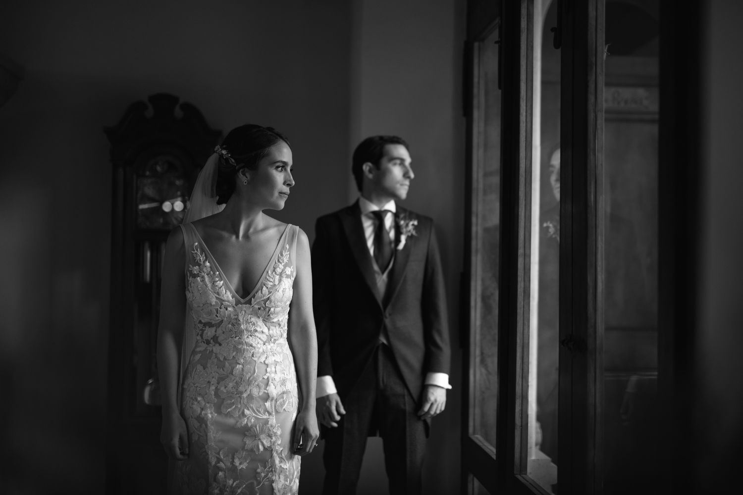 A black and white wedding photograph shows a couple in formal attire standing together by a window.