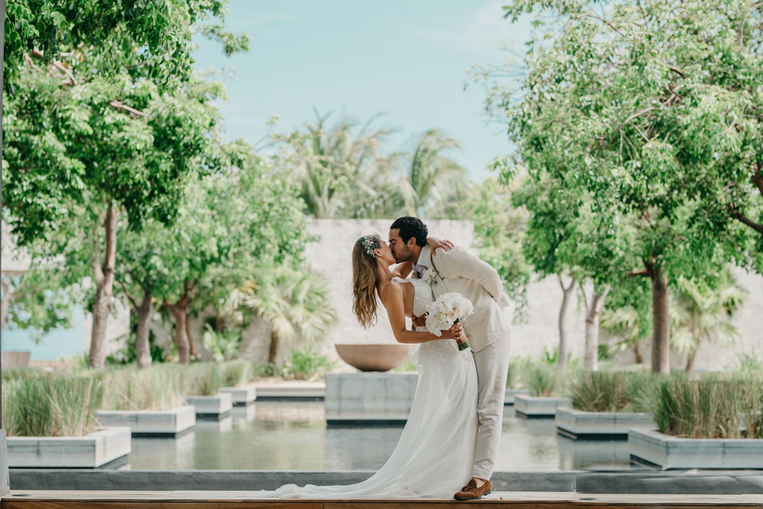 A couple in elegant white attire share a romantic moment near a reflecting pool surrounded by tropical palm trees.