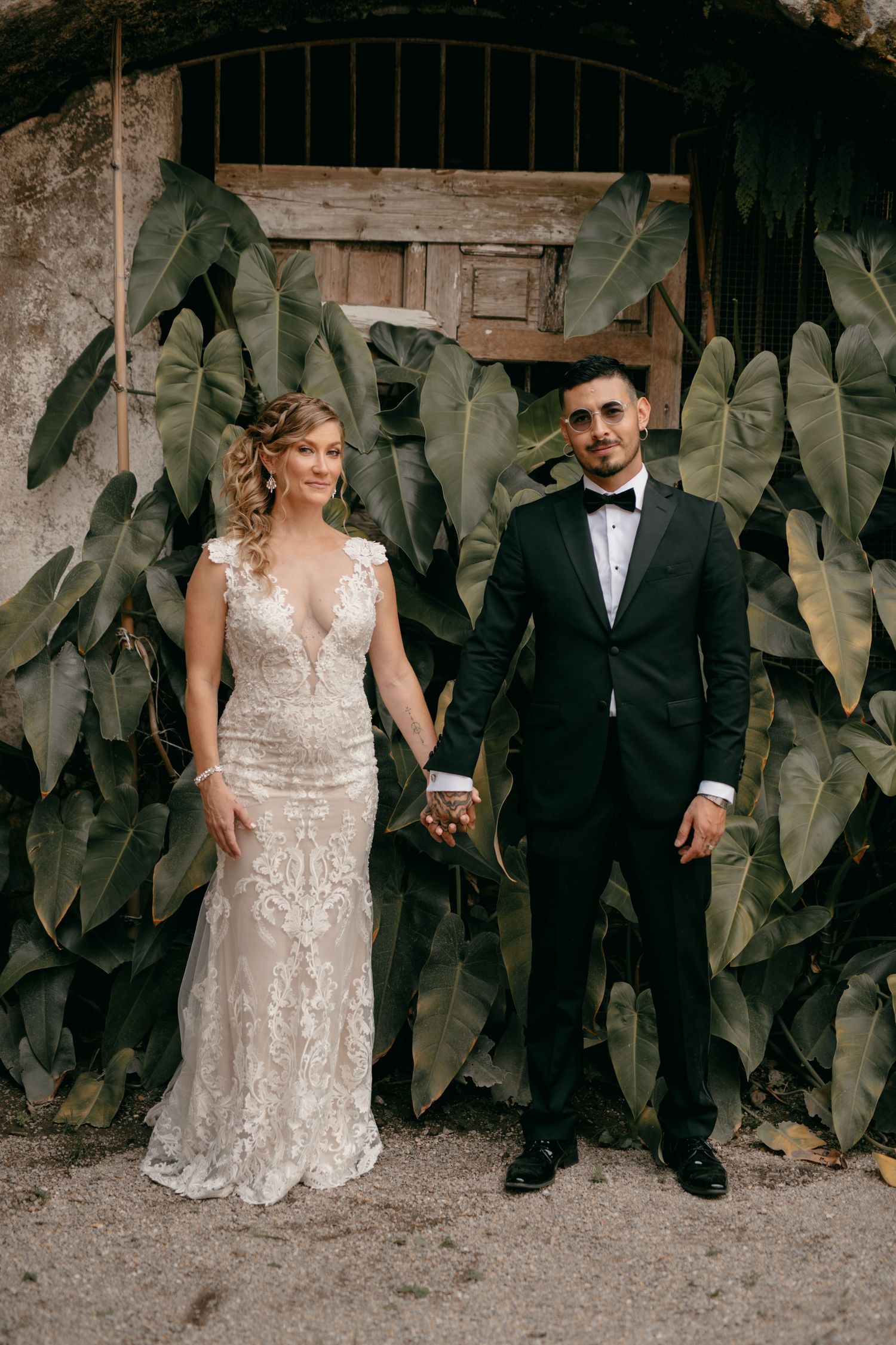 A newlywed couple poses in front of large tropical leaves, with bride in lace gown and groom in black tuxedo.
