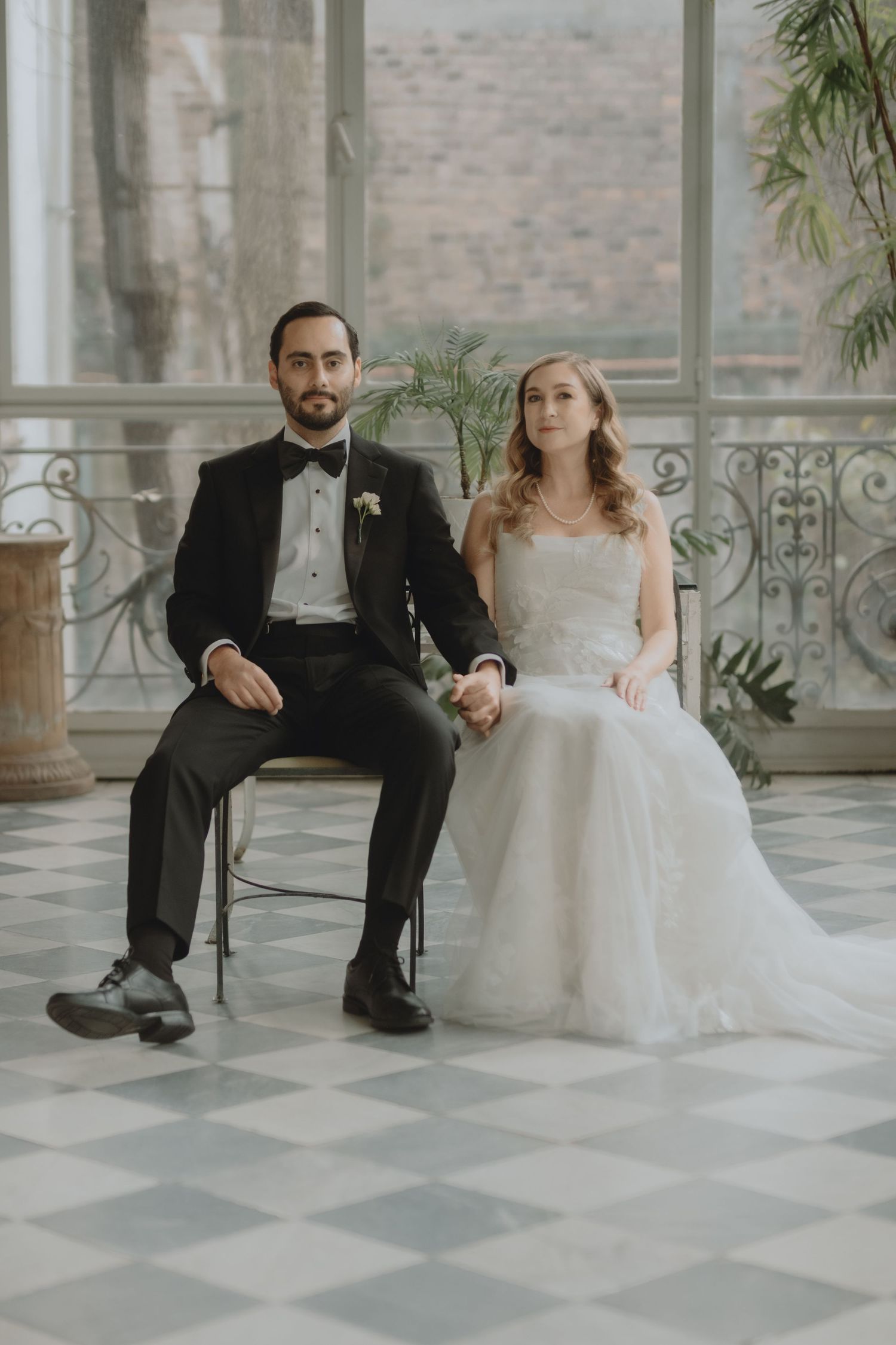 Wedding couple seated on chairs in sunlit room with checkered floor and vintage decor.