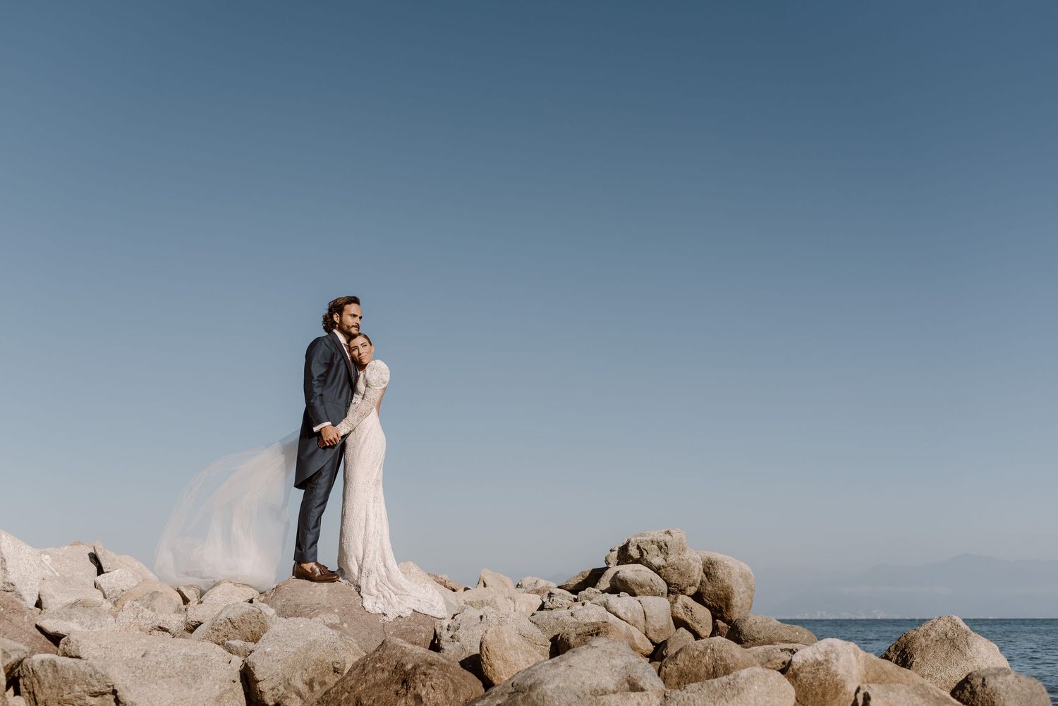 Dramatic coastal wedding portrait with couple standing on rocky shoreline against blue sky.