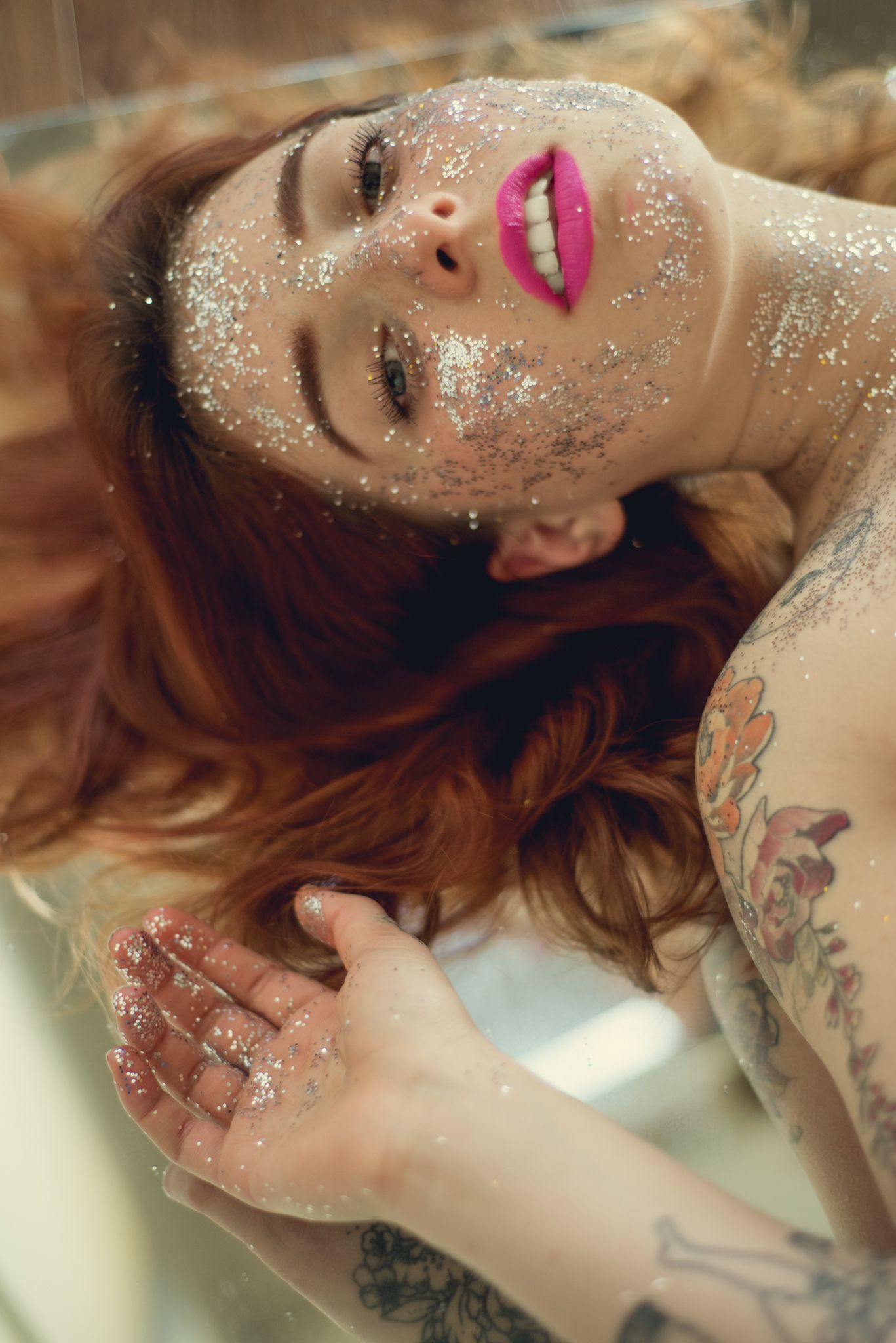 Ethereal beauty portrait with scattered glitter, visible tattoos and bright pink lipstick.