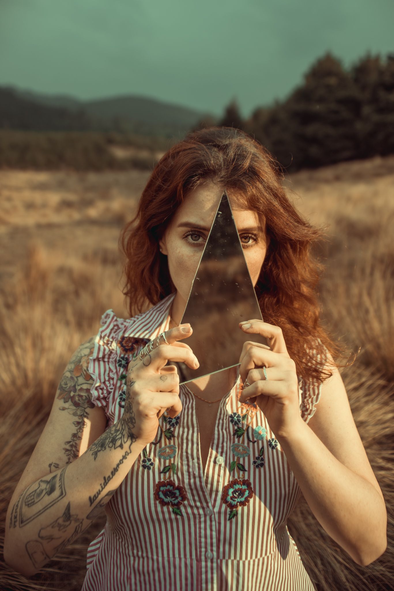 Person in vintage dress holds triangular mirror fragment against rural mountain landscape background.