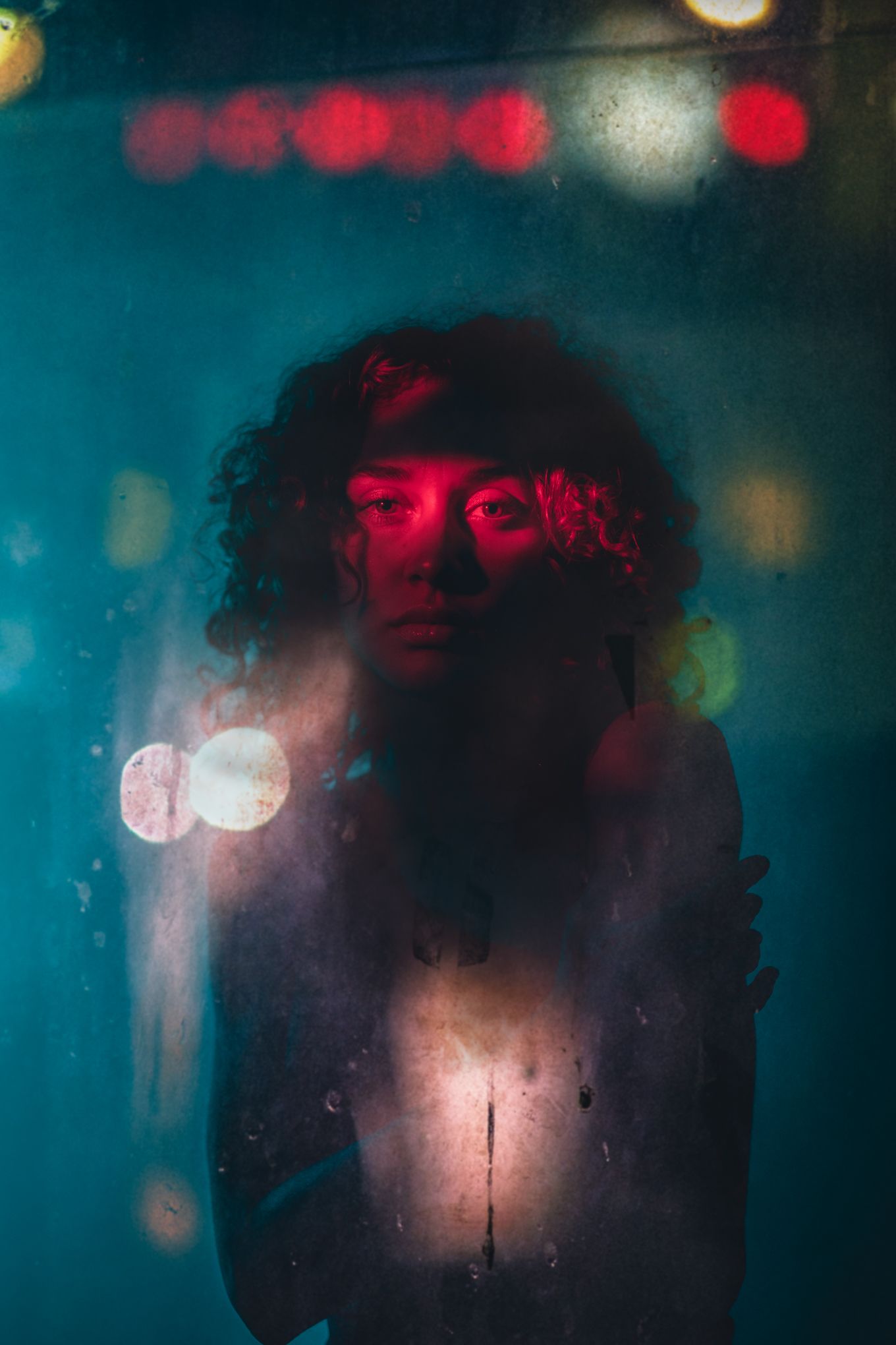 Moody portrait through glass with red lighting and bokeh effects in background.