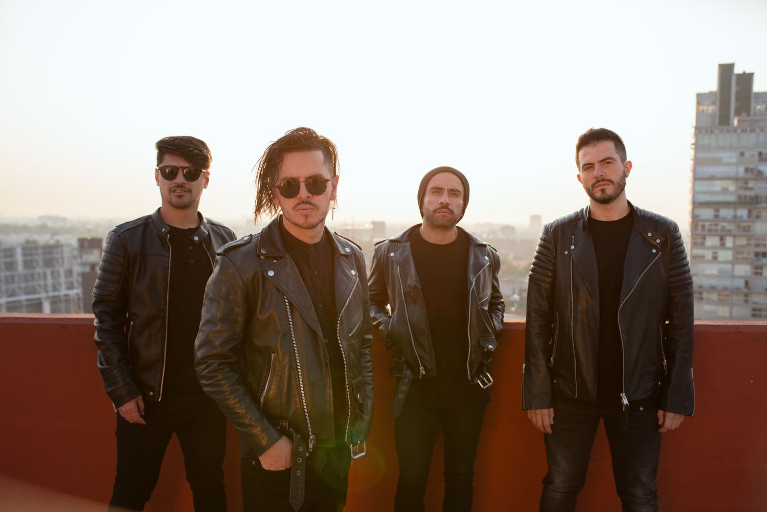 Four musicians pose together on a rooftop wearing black leather jackets against a sunset cityscape backdrop.