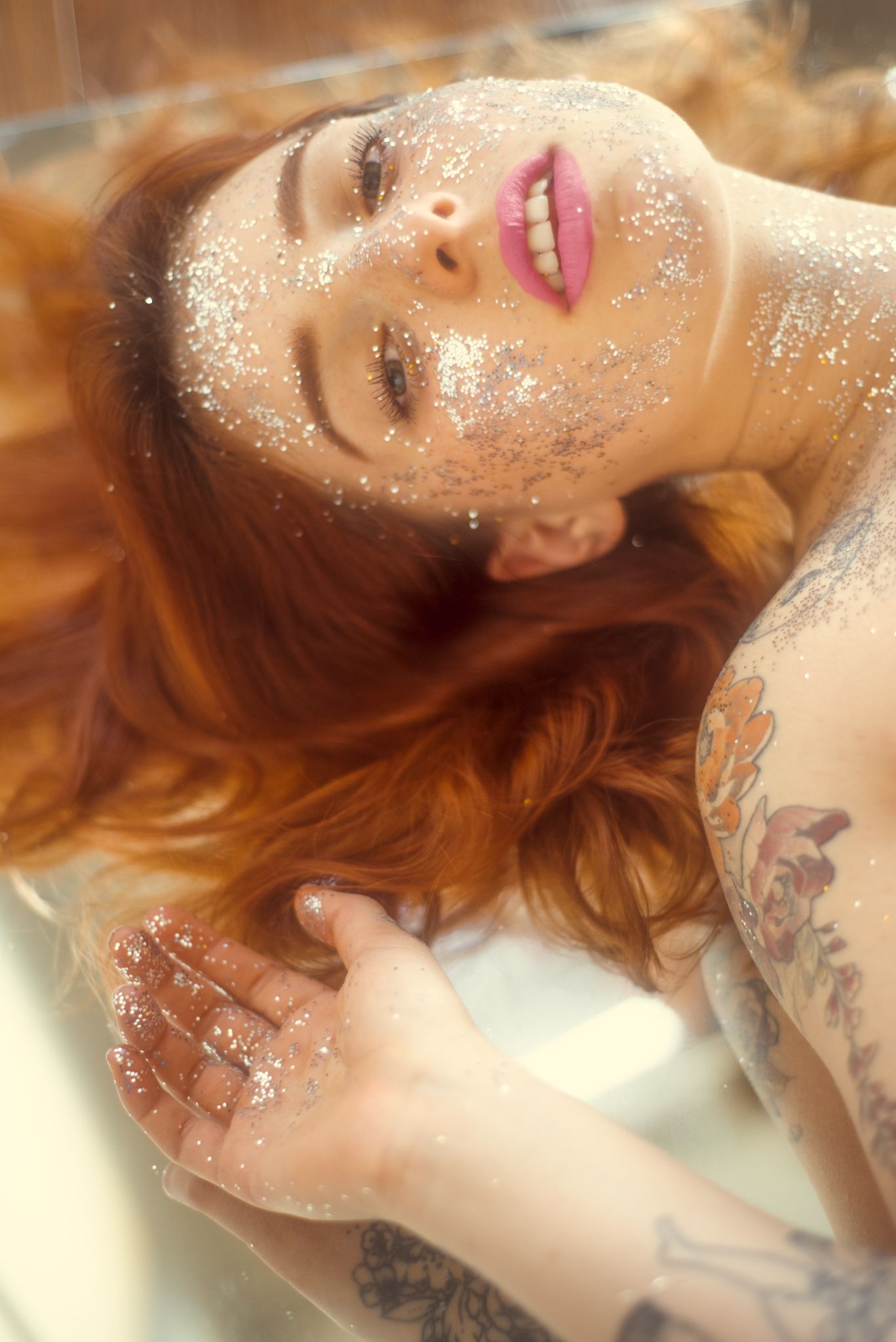 Artistic beauty shot with scattered glitter, tattoos and auburn hair in ethereal lighting.