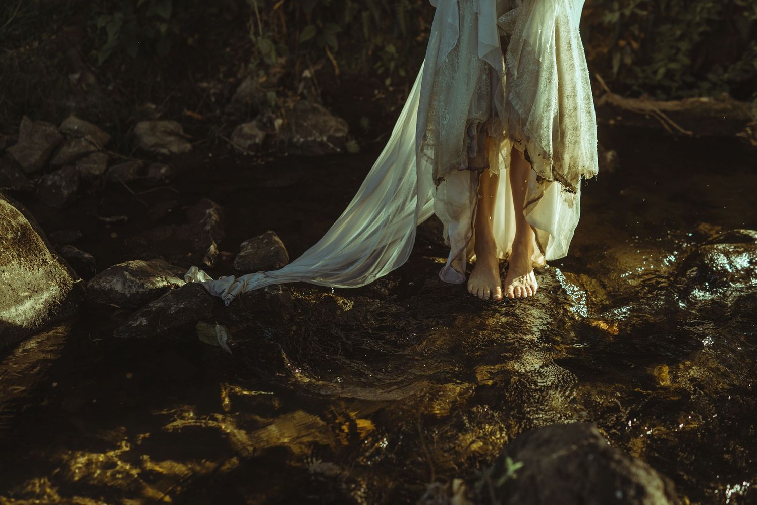 Ethereal white dress billows near bare feet standing on mossy rocks in shallow dark water at dusk.