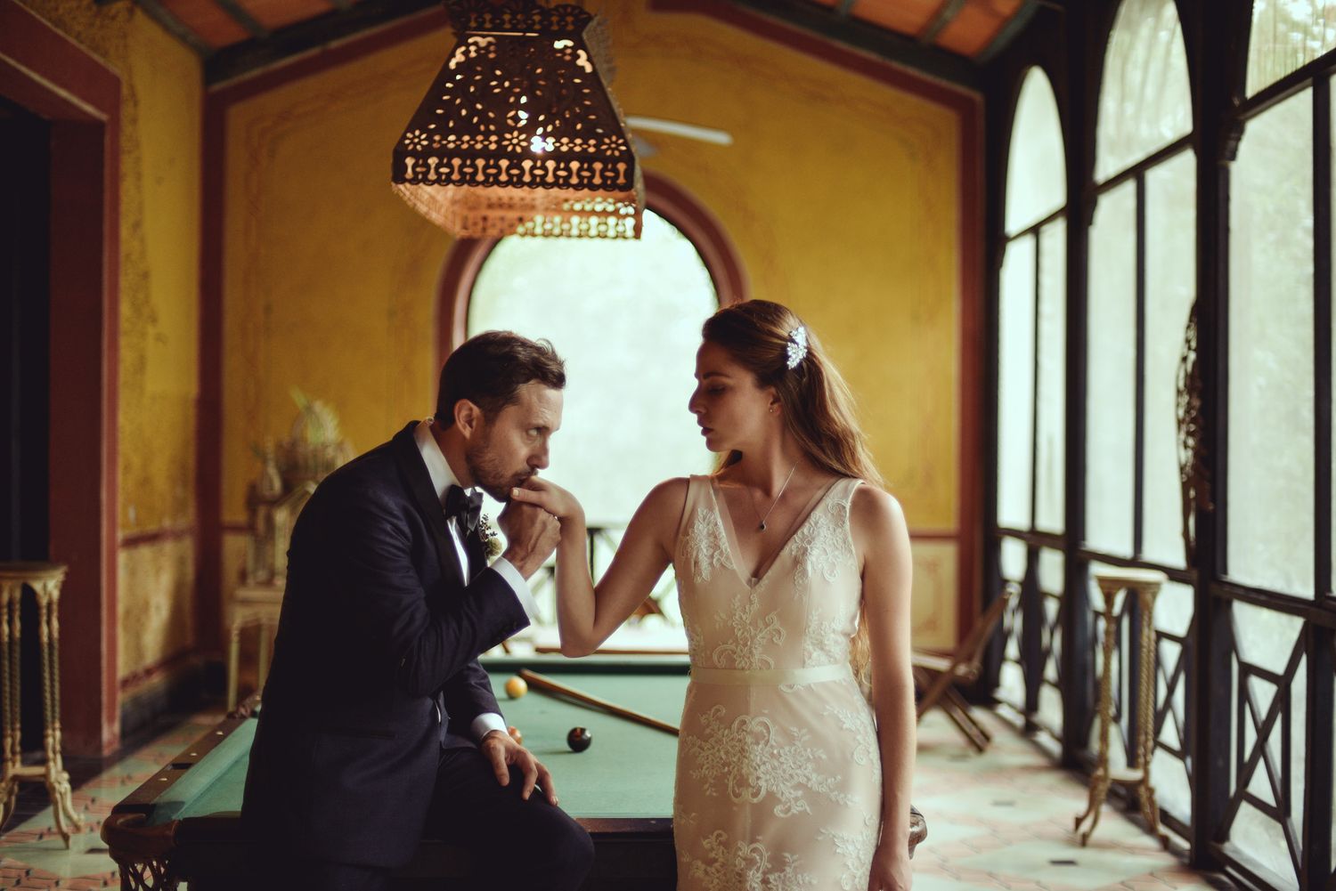 Elegant bride in lace gown and groom in tuxedo share romantic moment in vintage room with arched windows.