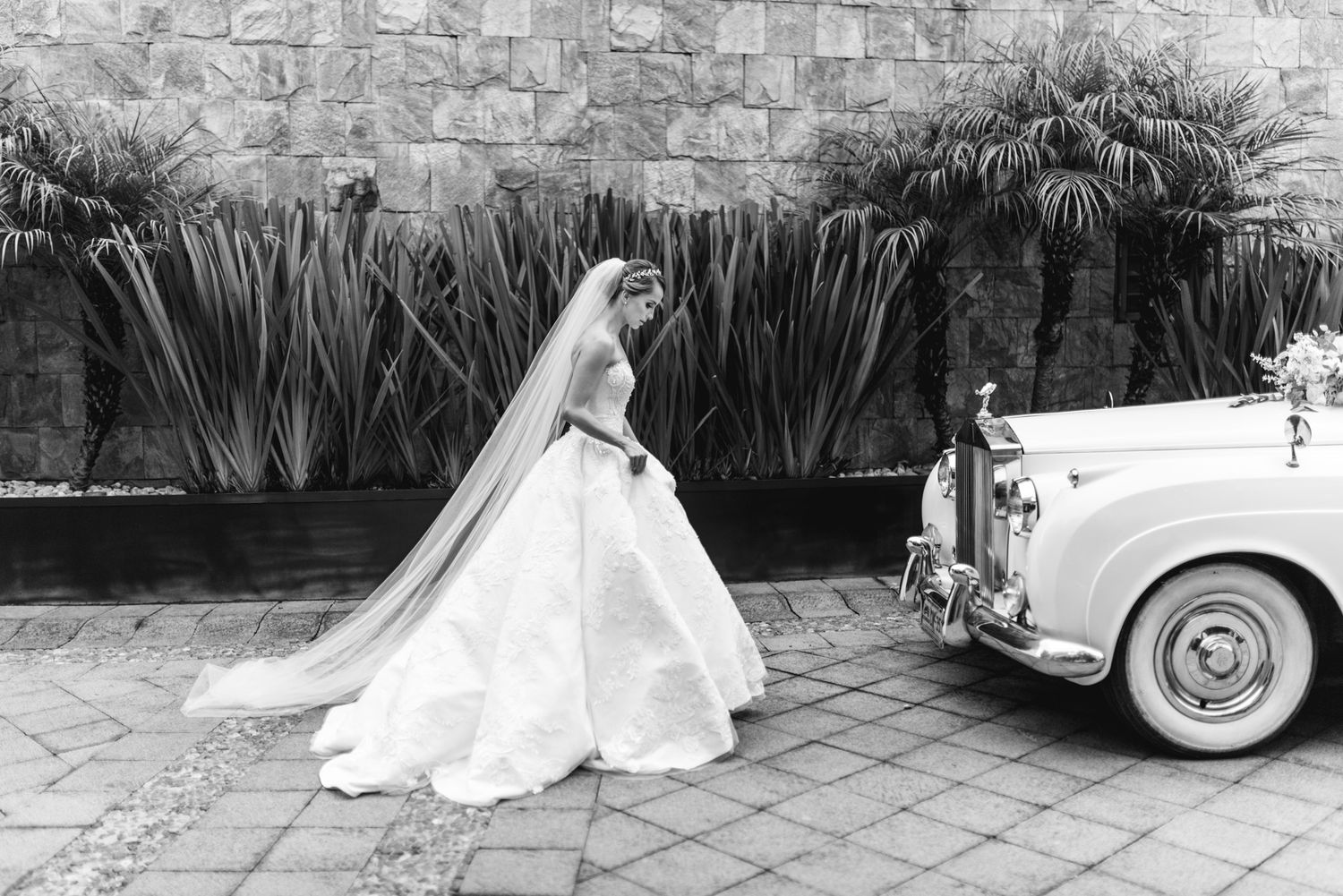 A wedding dress wearing person stands near a vintage Rolls Royce on a stone paved courtyard with foliage backdrop.
