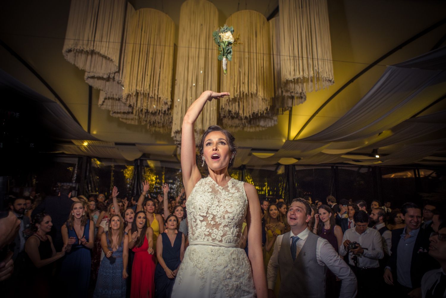 A bride in a white lace dress celebrating at her wedding reception while guests look on in a dimly lit ballroom.