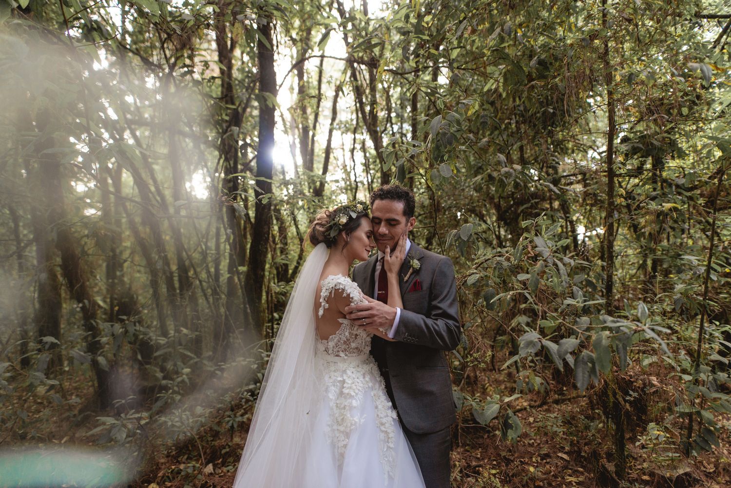 A romantic couple in wedding attire share an intimate moment in a sunlit forest with light rays streaming through trees.