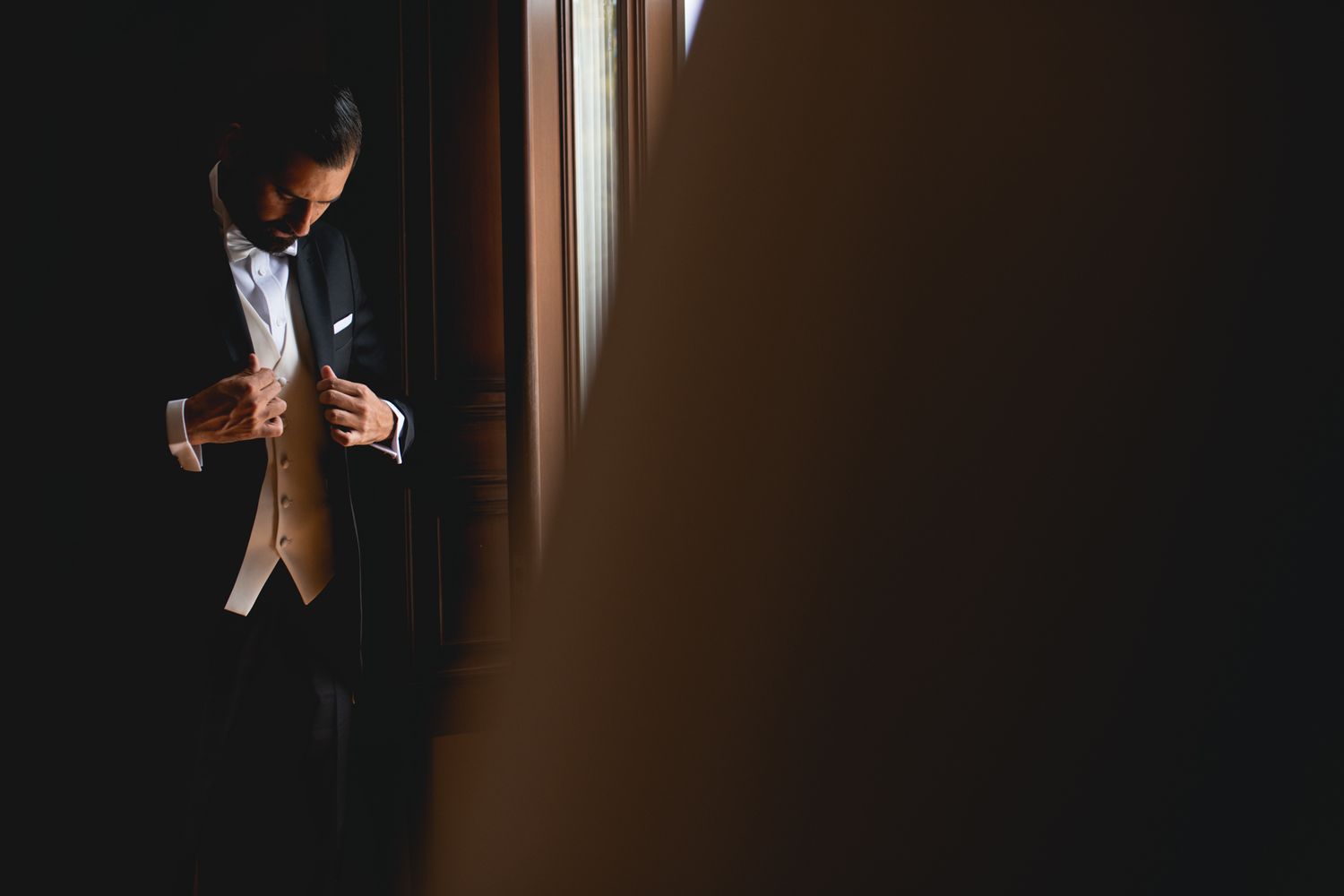 A figure in formal tuxedo attire adjusts their vest while standing in dramatic window lighting.
