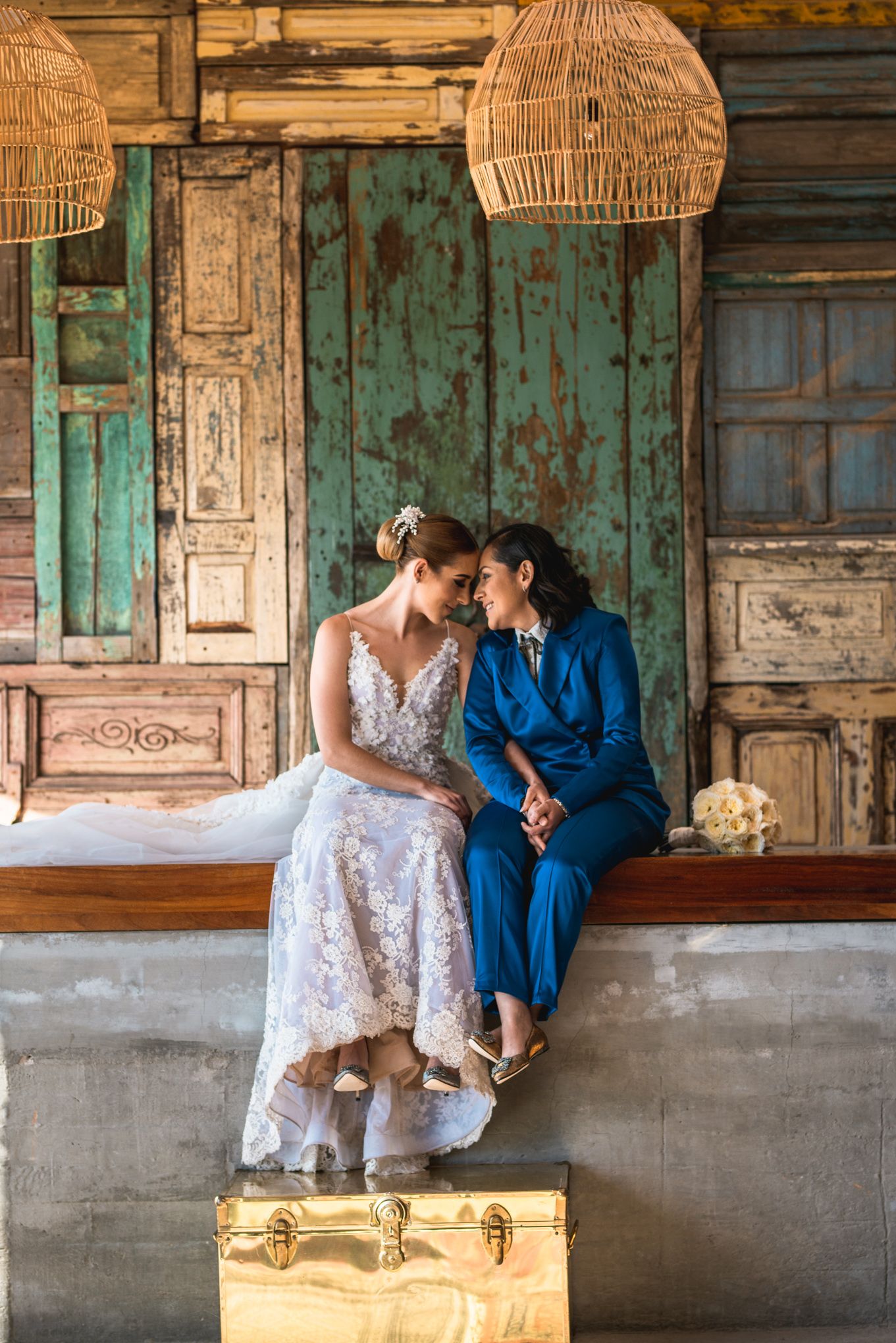 Two people in wedding attire sit together on a rustic wooden bench with woven pendant lights and vintage suitcase.