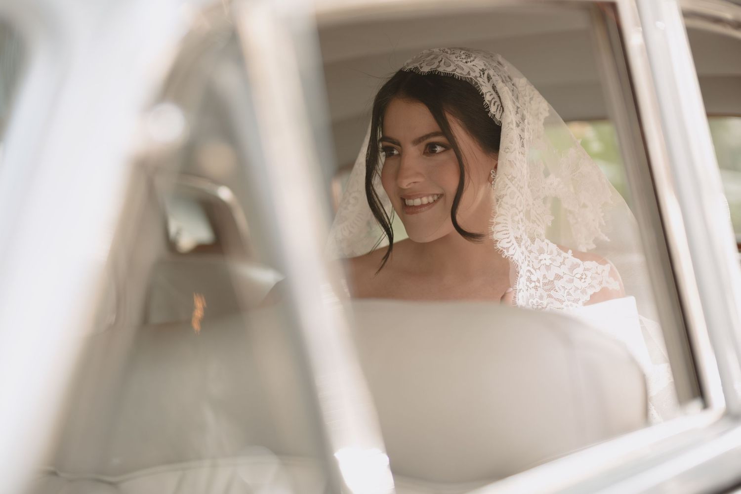 Beautiful bride with delicate lace veil smiles while sitting in vintage car on wedding day.