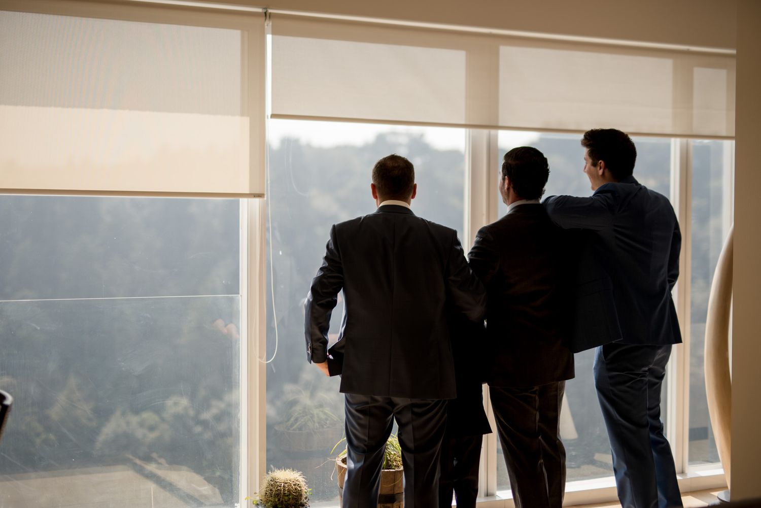 Three figures in suits standing by window overlooking scenic mountain view.